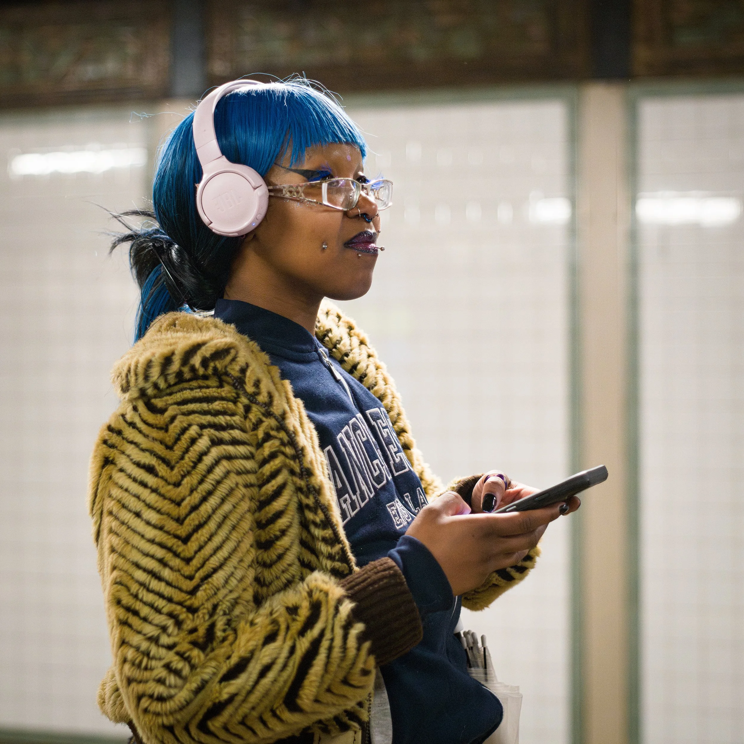 In New York Manhattan Subway, A young woman with blue hair, wearing pink headphones and glasses, is looking at her phone while standing indoors. She has a nose and lip piercings and is dressed in a fuzzy tiger-striped coat over a dark jacket.