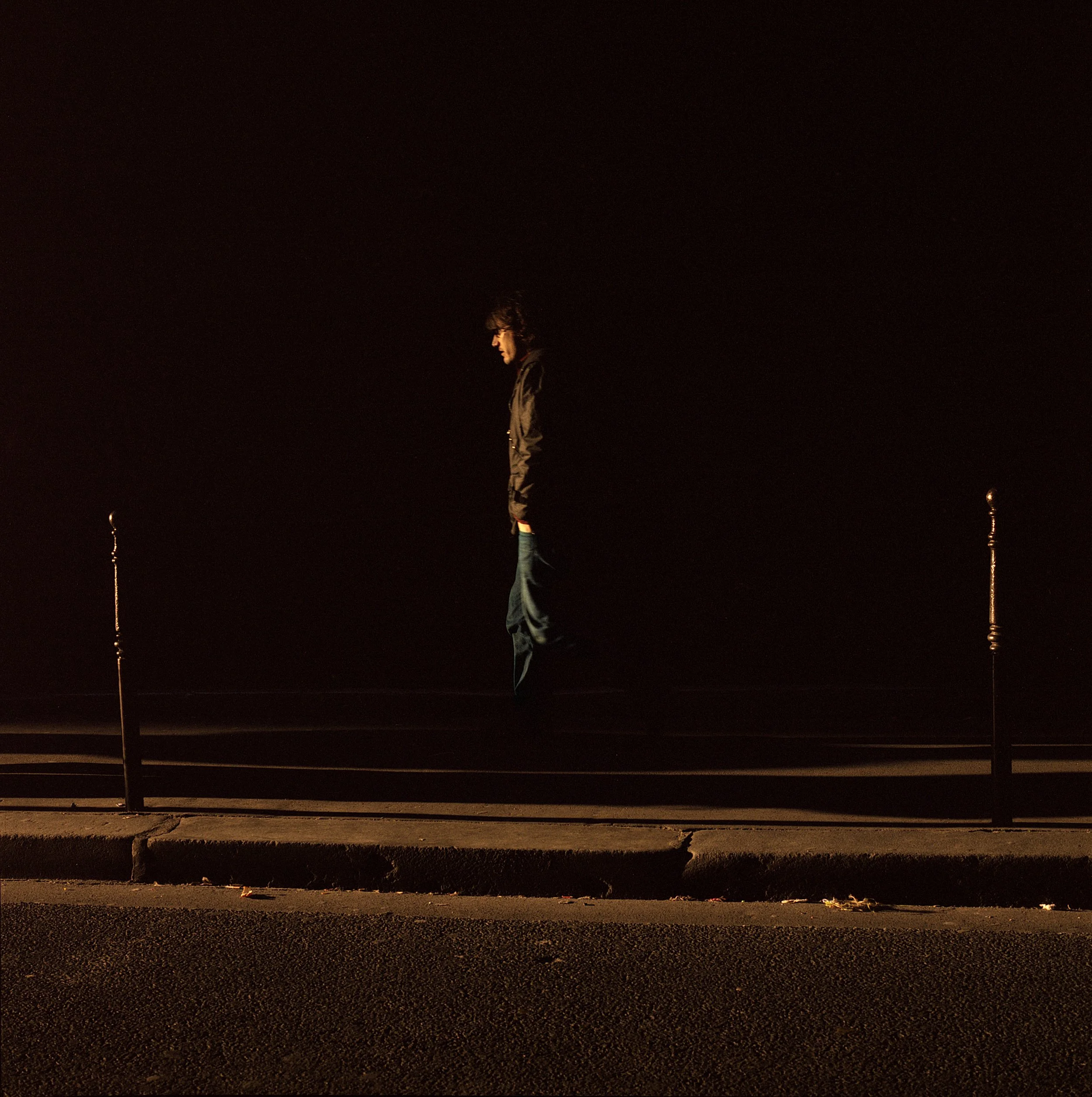 In Paris, France, A person standing alone at night, illuminated softly from the side, against a pitch-black background, with a railing and streetlamp visible in the foreground.
