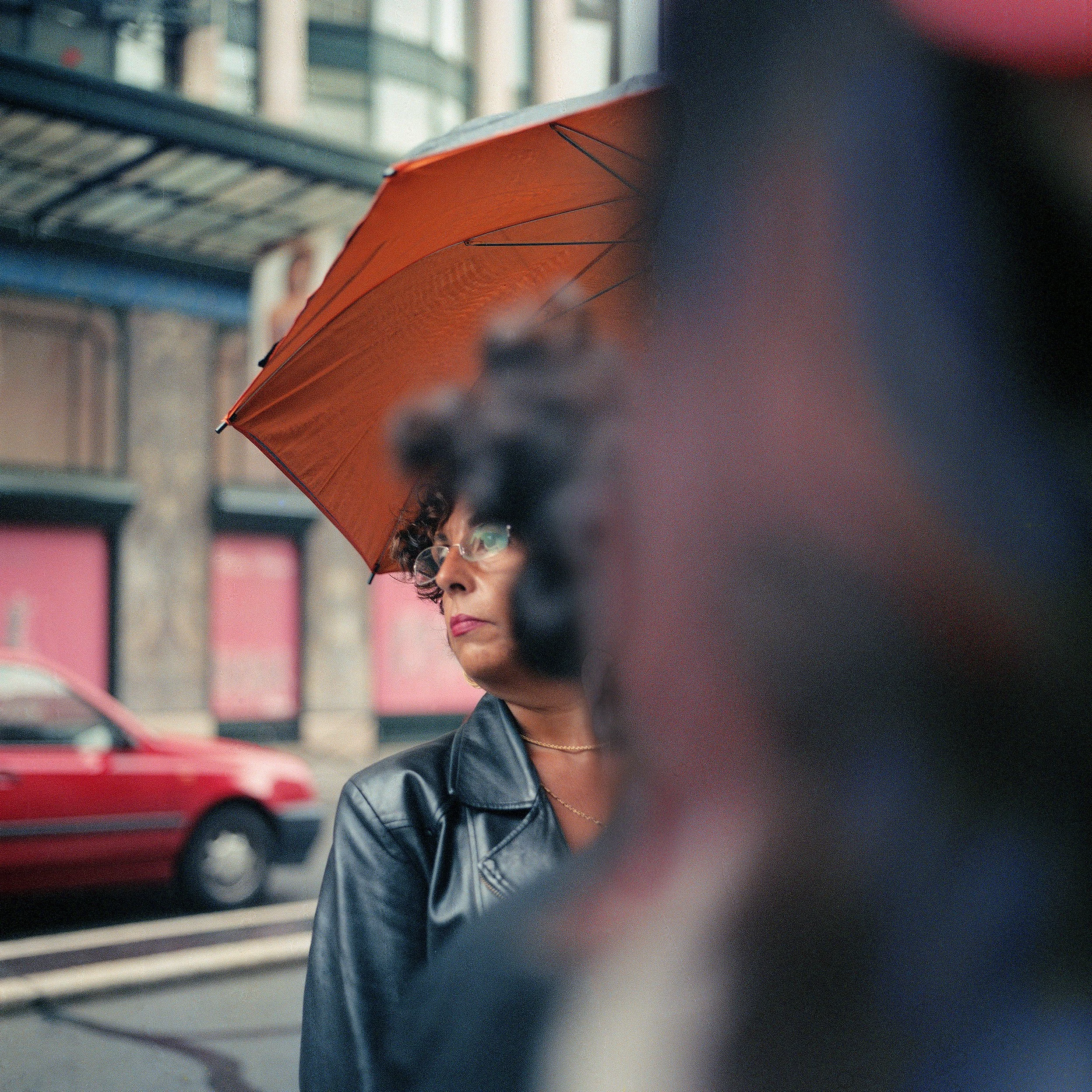 In Paris, France, Woman with glasses under an orange umbrella on a city street, wearing a black leather jacket.