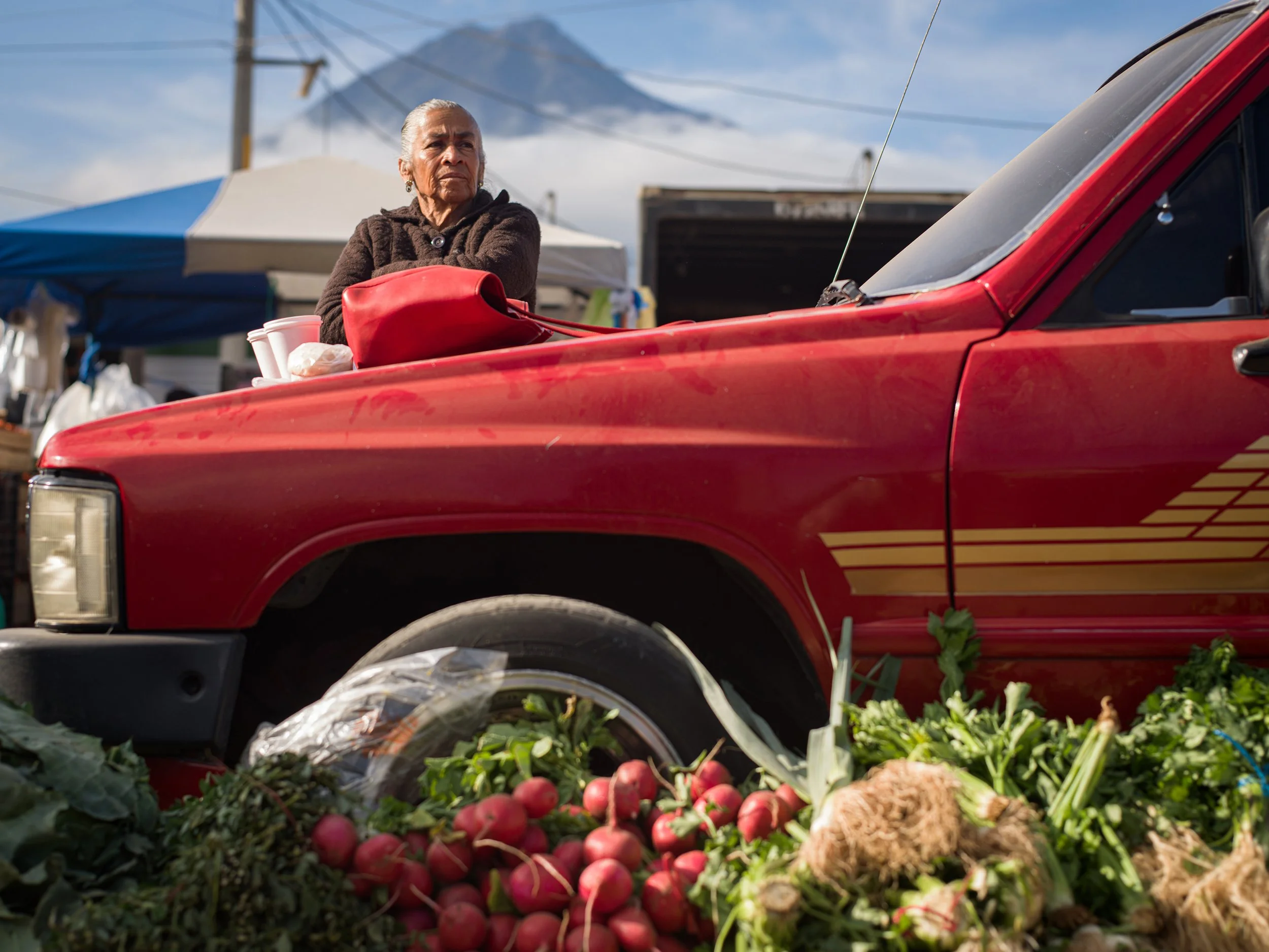In Antigua market, Guatemala, An elderly woman at an outdoor market standing behind a red vehicle with fresh vegetables and radishes in front.