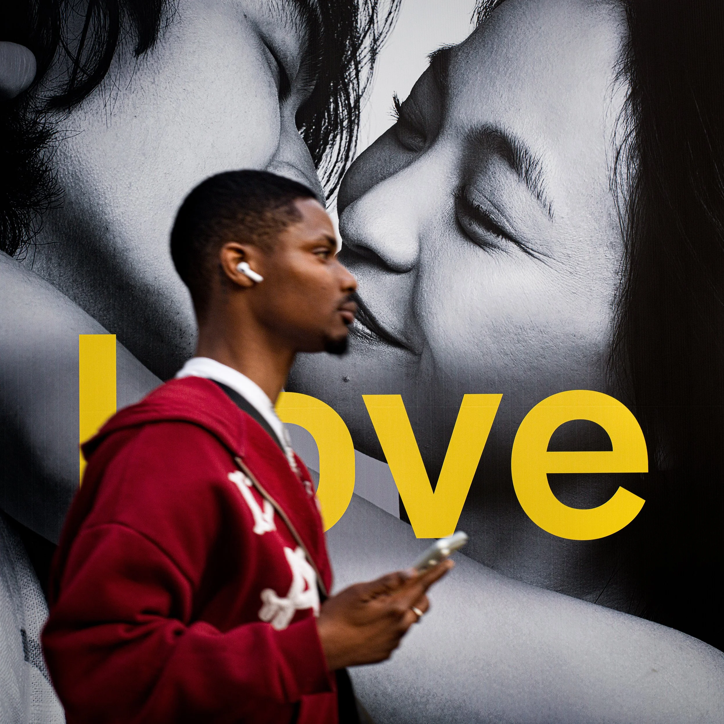 In New York Manhattan, A young man in a red hoodie and white earbud, holding a smartphone, walking past a billboard with a black and white photo of two women close to kissing, with the word 'Love' in yellow.
