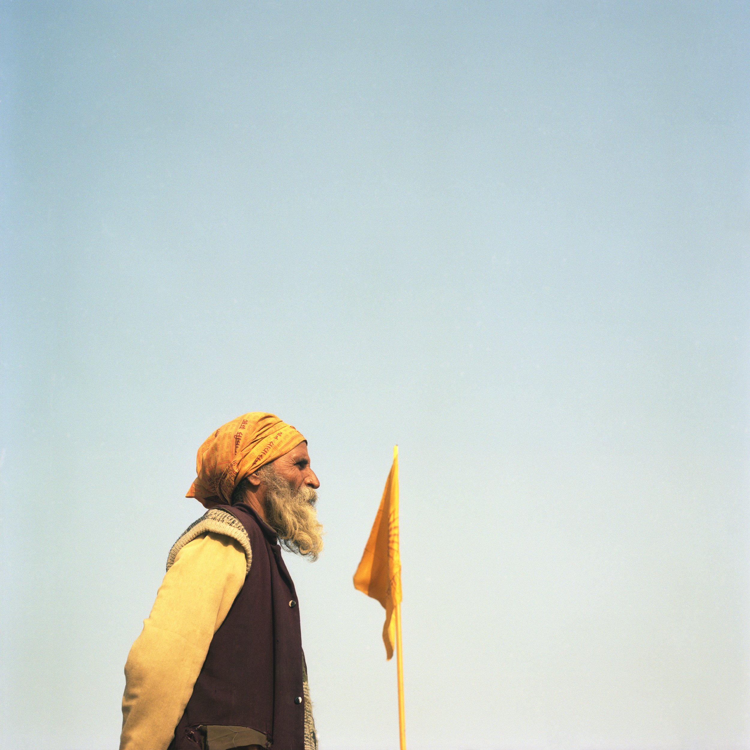 A man with a beard wearing a beige headscarf and a brown vest standing outdoors next to a yellow flag against a clear blue sky.