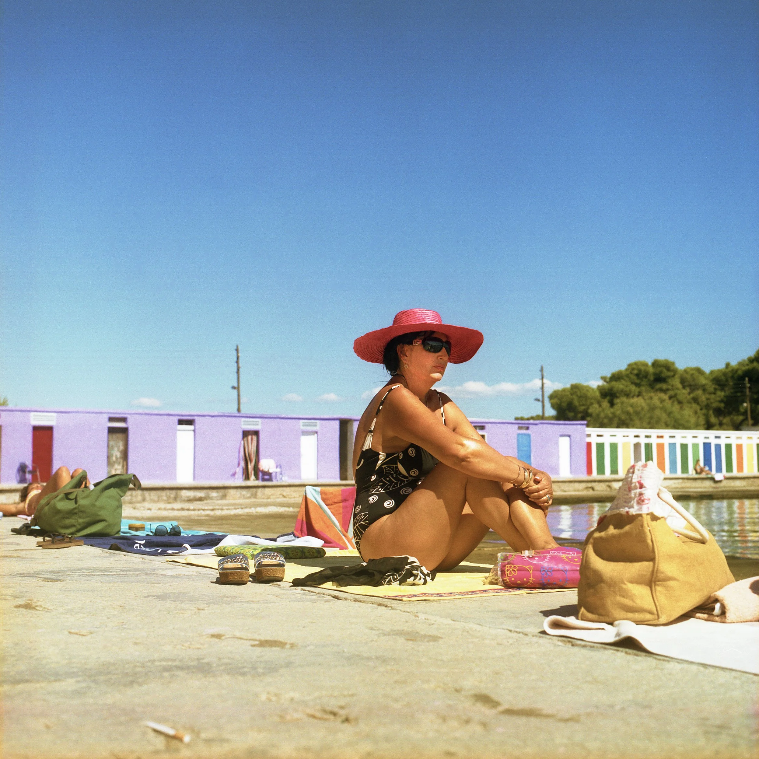Woman sitting on a towel by the lake, wearing a wide-brimmed pink hat and sunglasses, with colorful bags and shoes nearby, and a purple building with a colorful mural in the background.