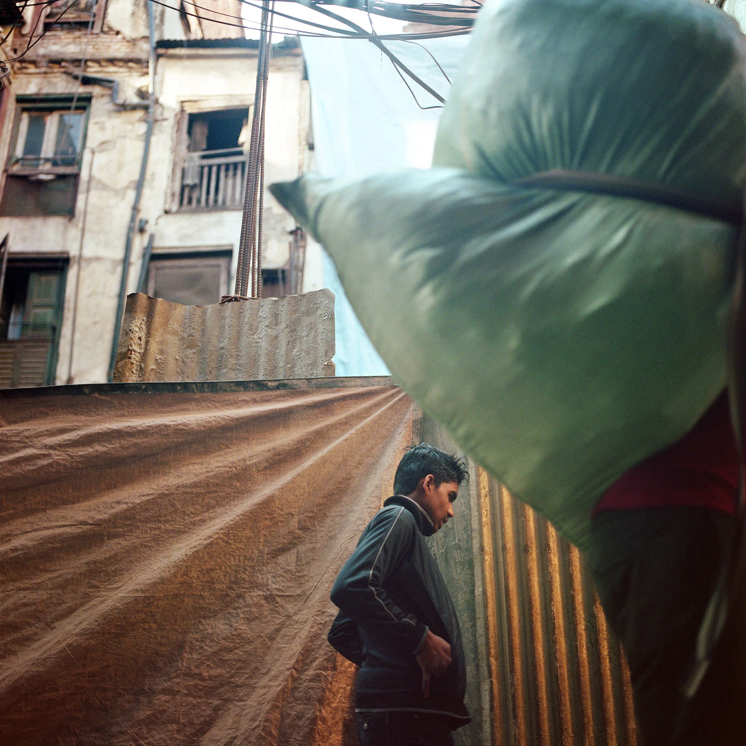 In Kathmandu, A young man standing with his hands in his pockets in a narrow outdoor alleyway between old, weathered apartment buildings with visible balconies and windows. Part of a large green bag or tarp is visible in the foreground.