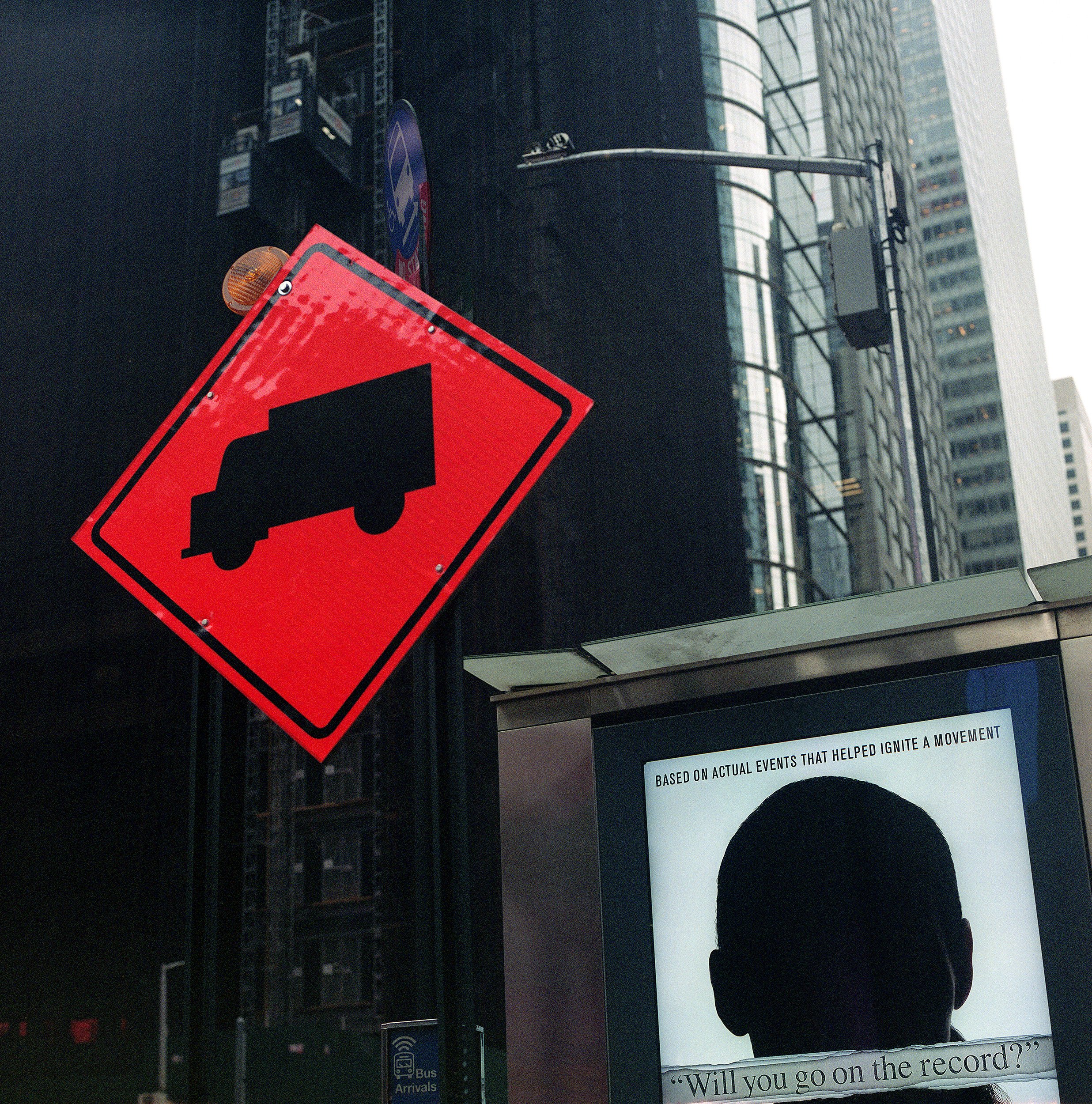 In New York Manhattan, an Urban street scene featuring a red warning sign with a truck icon, digital billboard displaying a silhouette of a person's head, and tall buildings in the background.
