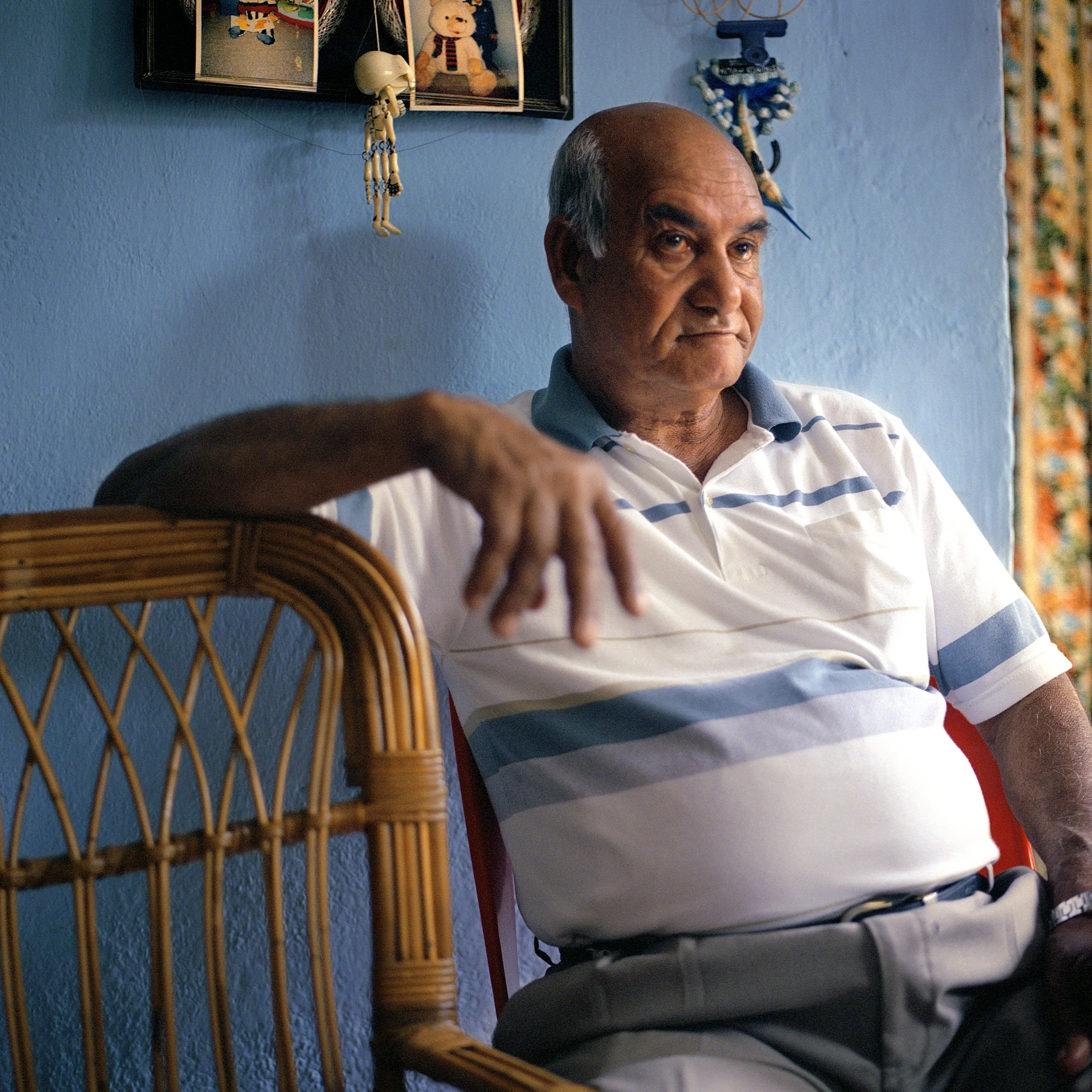 In Mauritius, Trou d'Eau Douce, An elderly man sitting in a room with blue walls, wearing a striped polo shirt, with framed photographs and decorative items on the wall behind him.