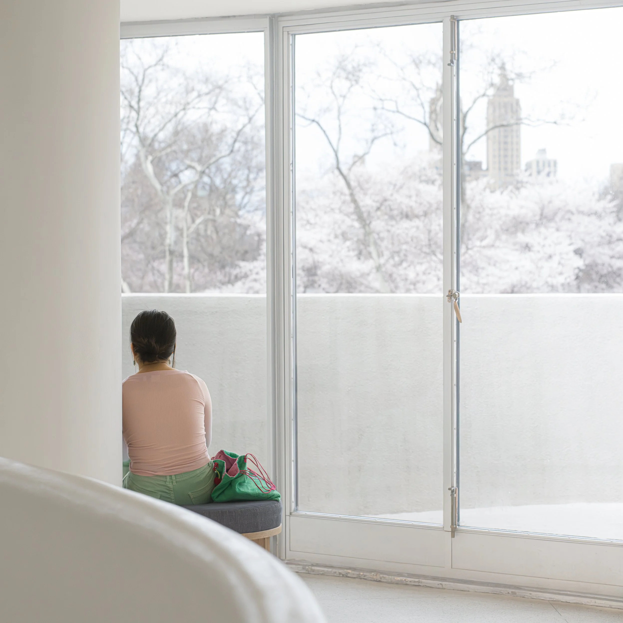 In New York Manhattan, Guggenheim Museum,  A woman sitting on a bench in a brightly lit room, looking out a large glass window at snow-covered trees and buildings.