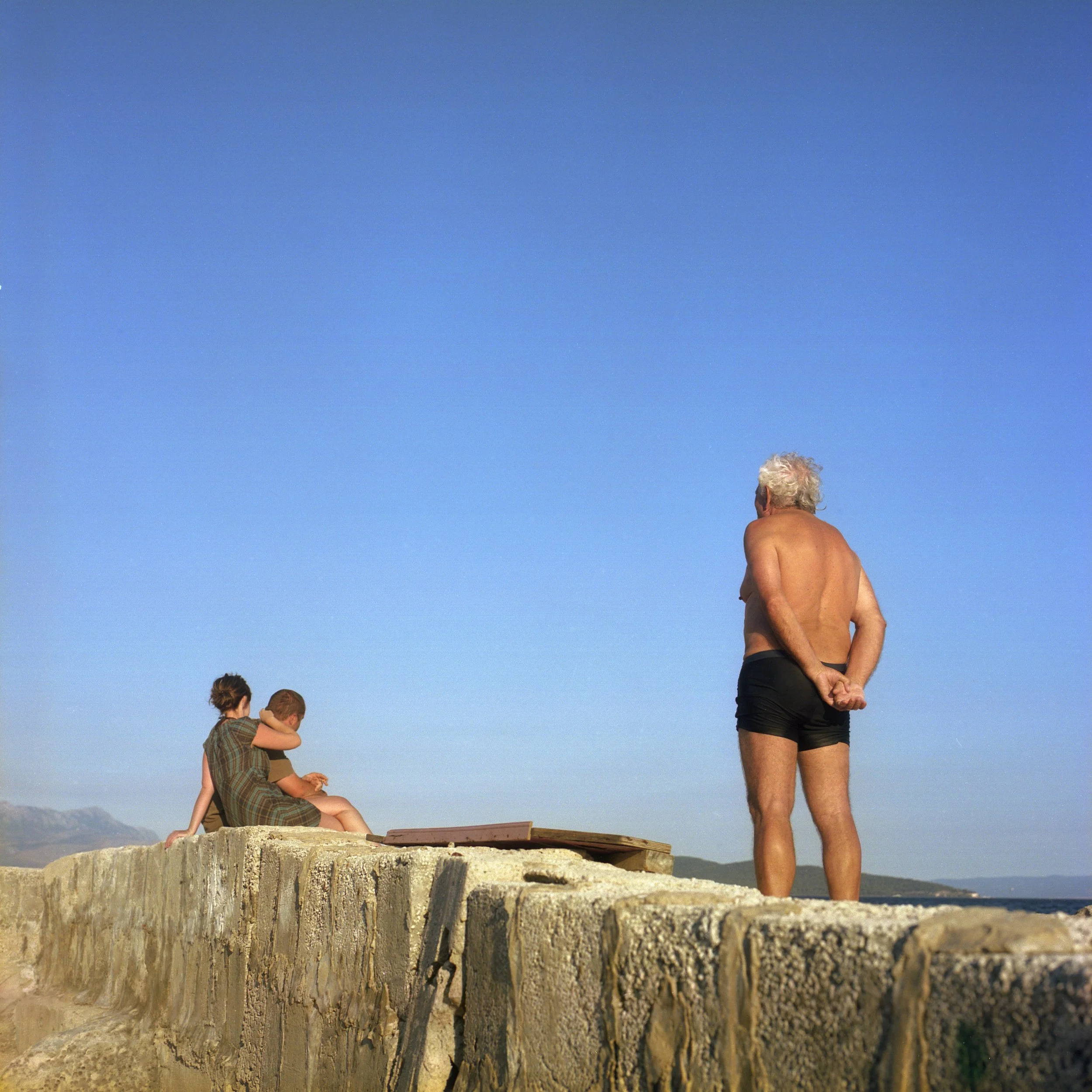 An elderly man in black shorts stands on a stone wall looking out toward the ocean, while two young people sit on the wall nearby, one embracing the other, with a mountainous landscape in the distance under a clear blue sky.