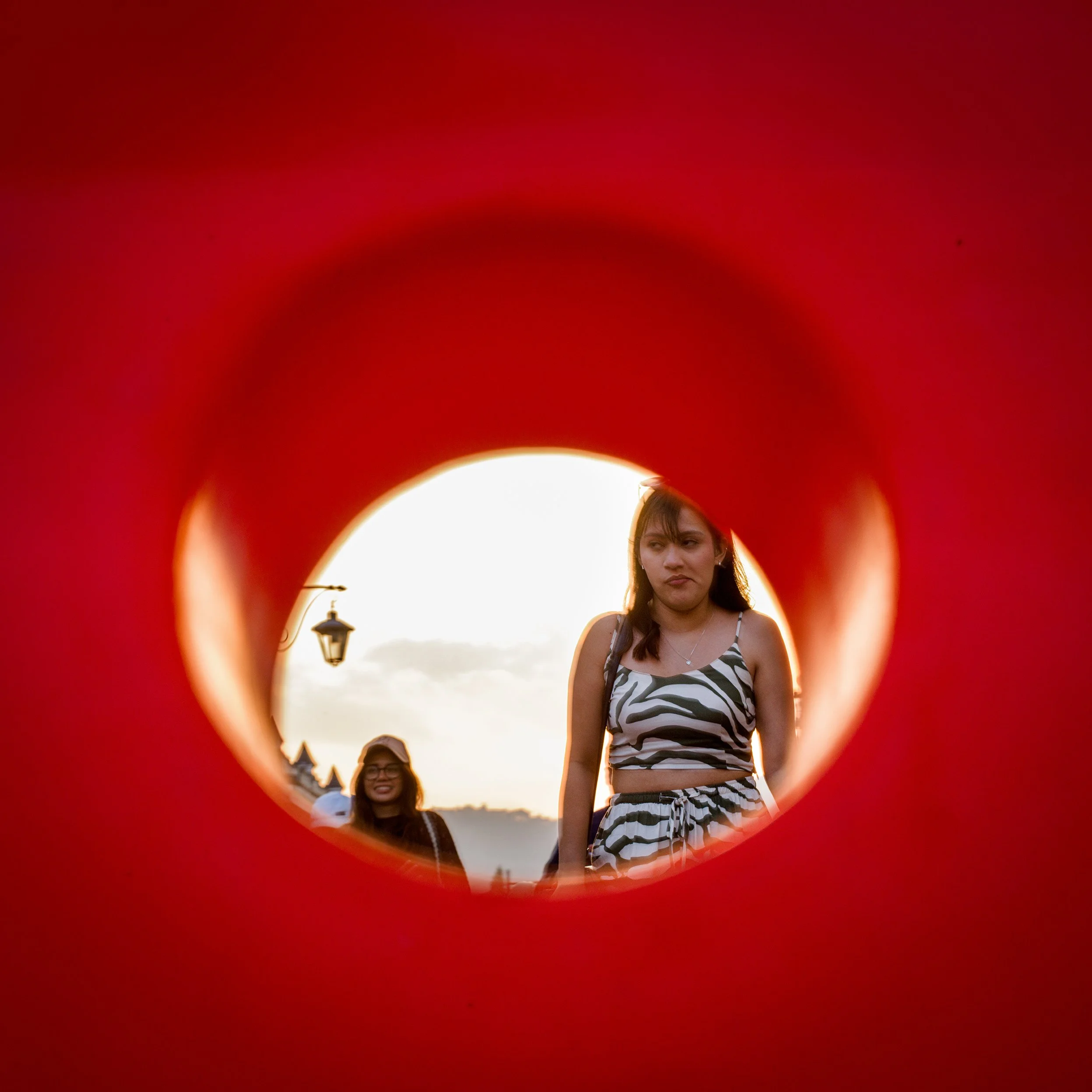 In Antigua, Guatemala, Two women seen through a small opening in a red structure, with one woman in a striped dress with an uncertain expression, and a smiling woman in glasses and dark clothing in the background, outdoors during sunset.