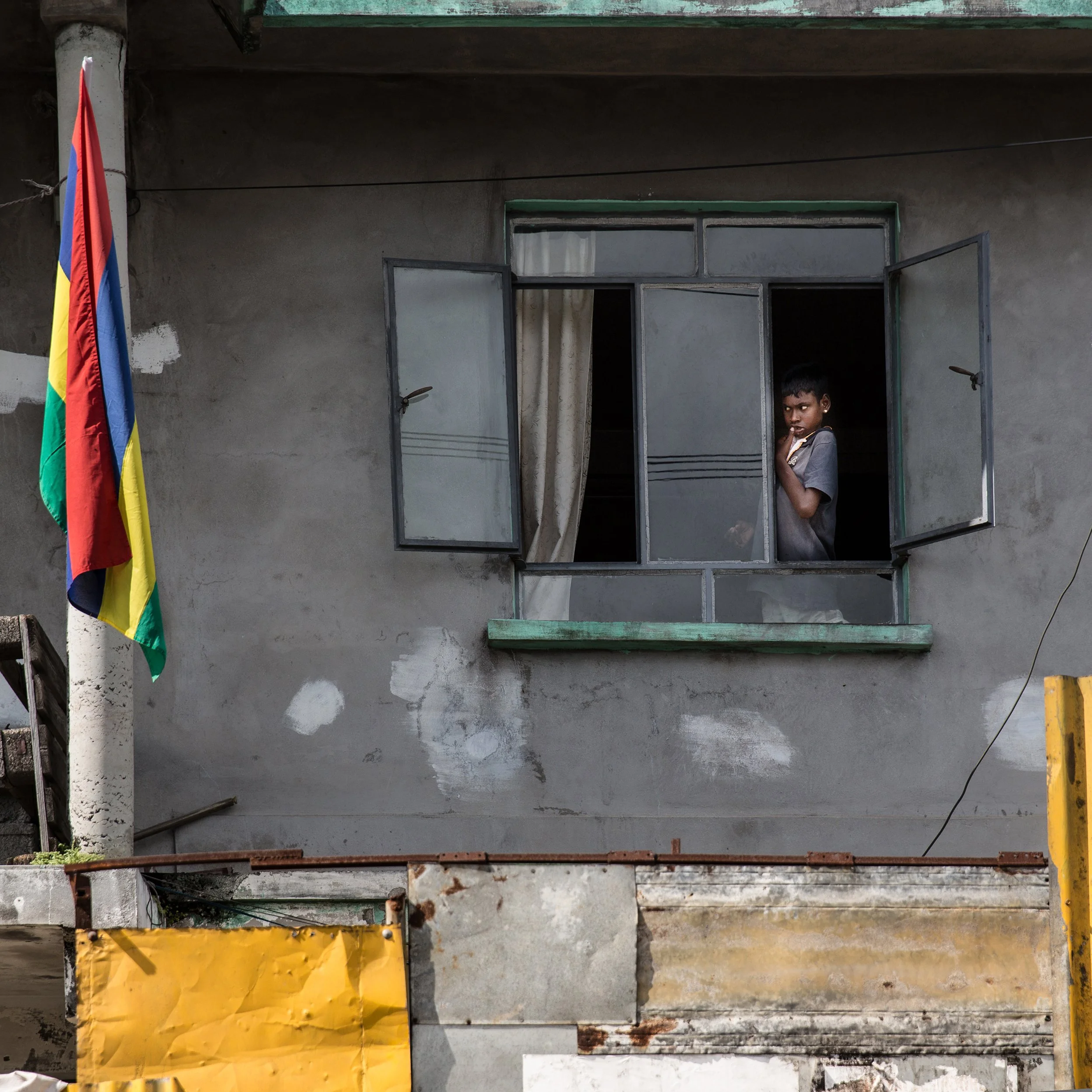 IN Mauritius, A young boy looking out of an open window on an unfinished gray concrete building with a Mauritian flag hanging on the left side.