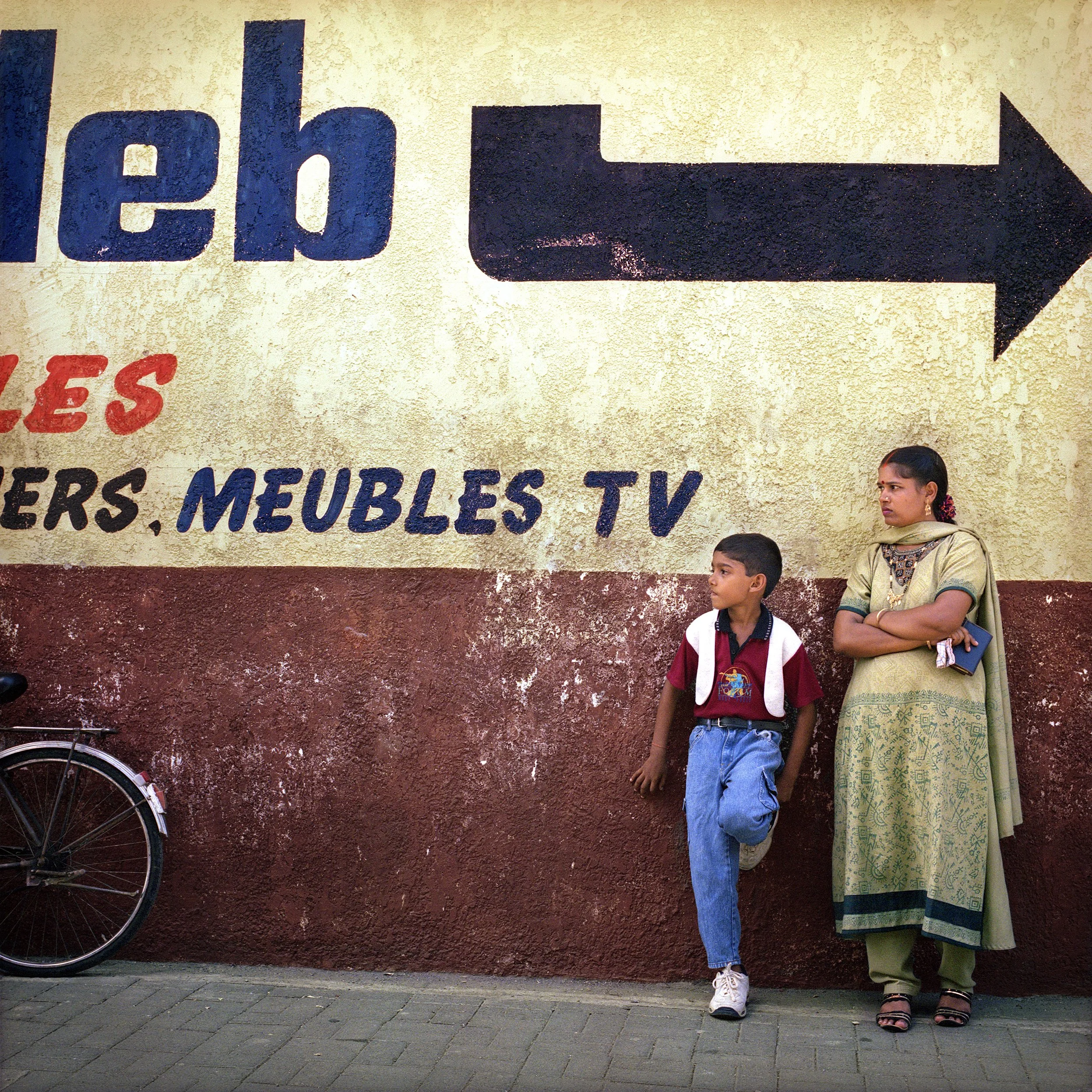 In Mauritius, Flack, A woman and a boy standing against a wall with a large painted advertisement, partially obscured, with an arrow pointing right. The woman is wearing traditional Indian clothing, the boy is in school uniform