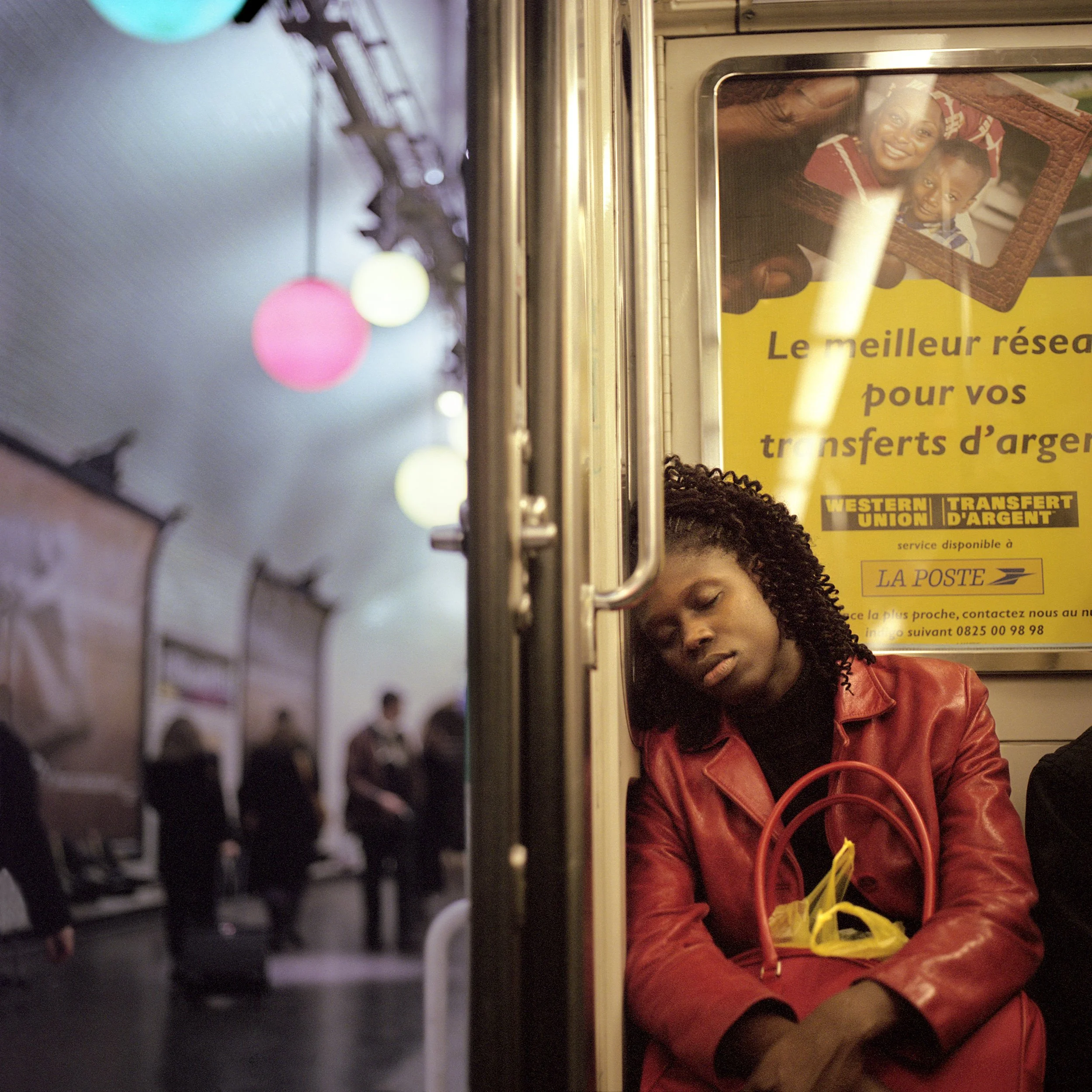 In Paris Subway, A woman with curly hair and a young boy with short hair sitting together on a train or bus, with a yellow advertisement poster behind them, and multicolored string lights in the foreground.