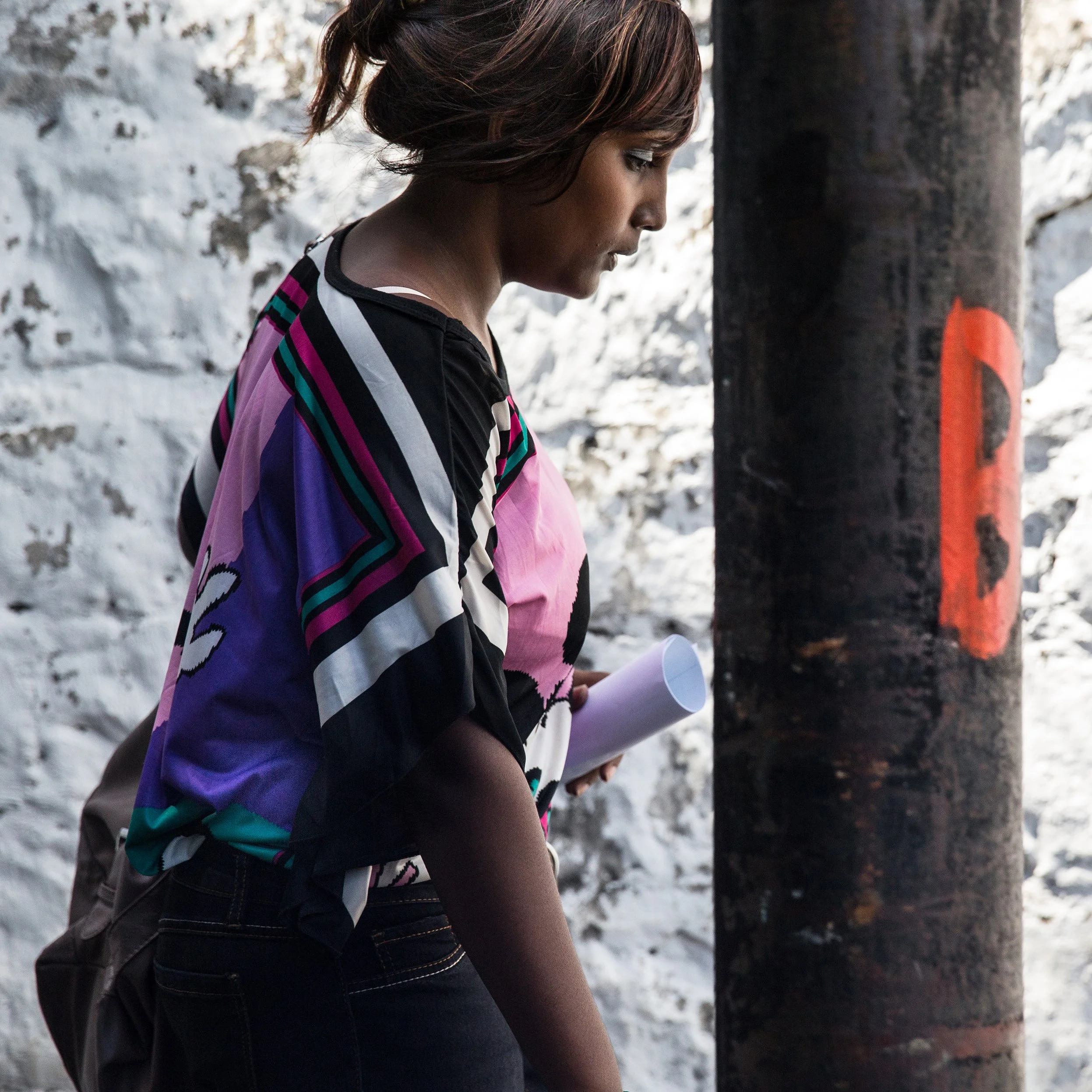 In Mauritius, Port Louis A woman with short brown hair, wearing a colorful striped jacket and black jeans, is walking in front of a white stone wall