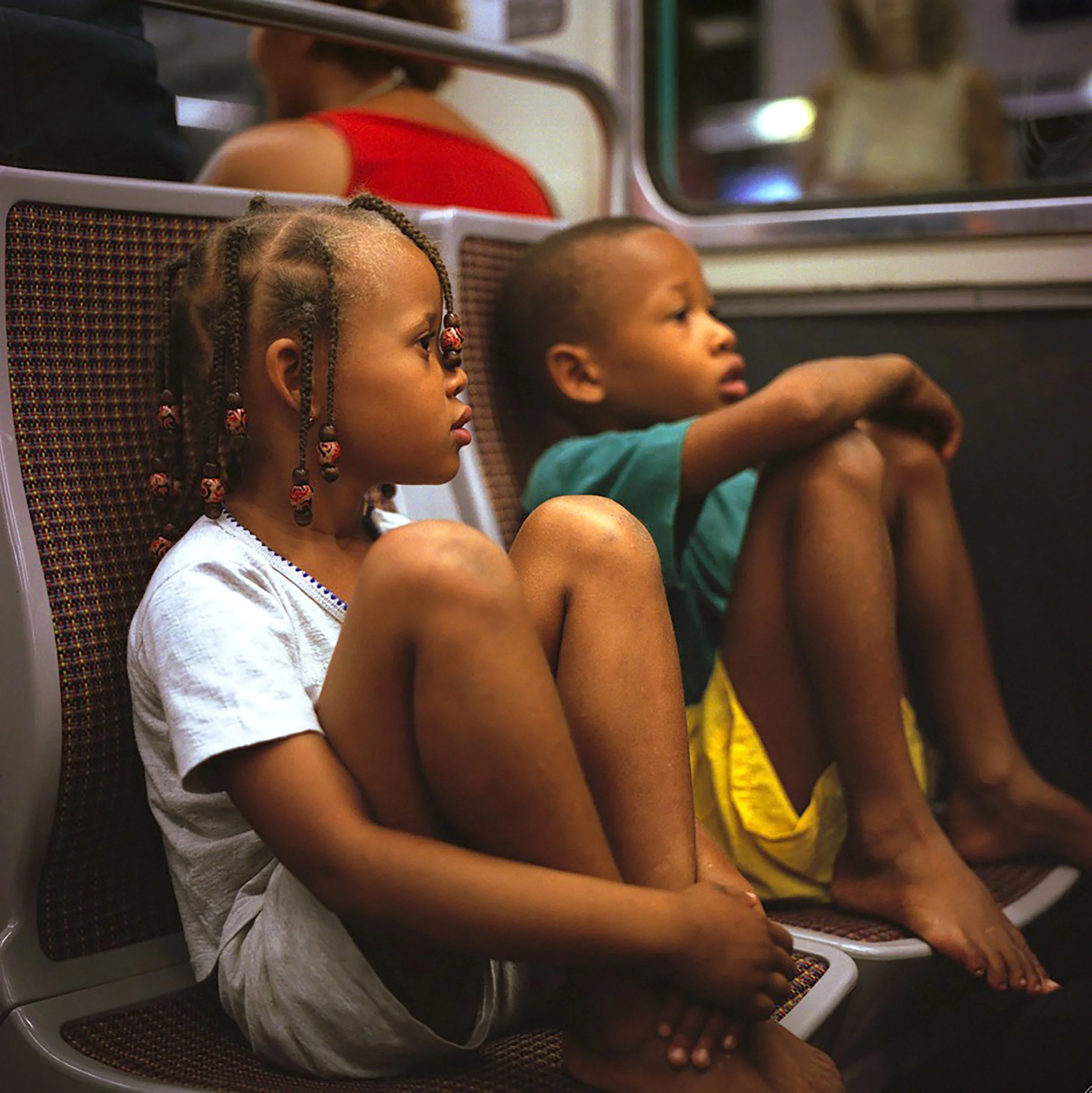 In Paris Subway, Two young children with braided hair, sitting side by side in what appears to be a subway or train, looking forward; the girl wears a white shirt, and the boy wears a green shirt.