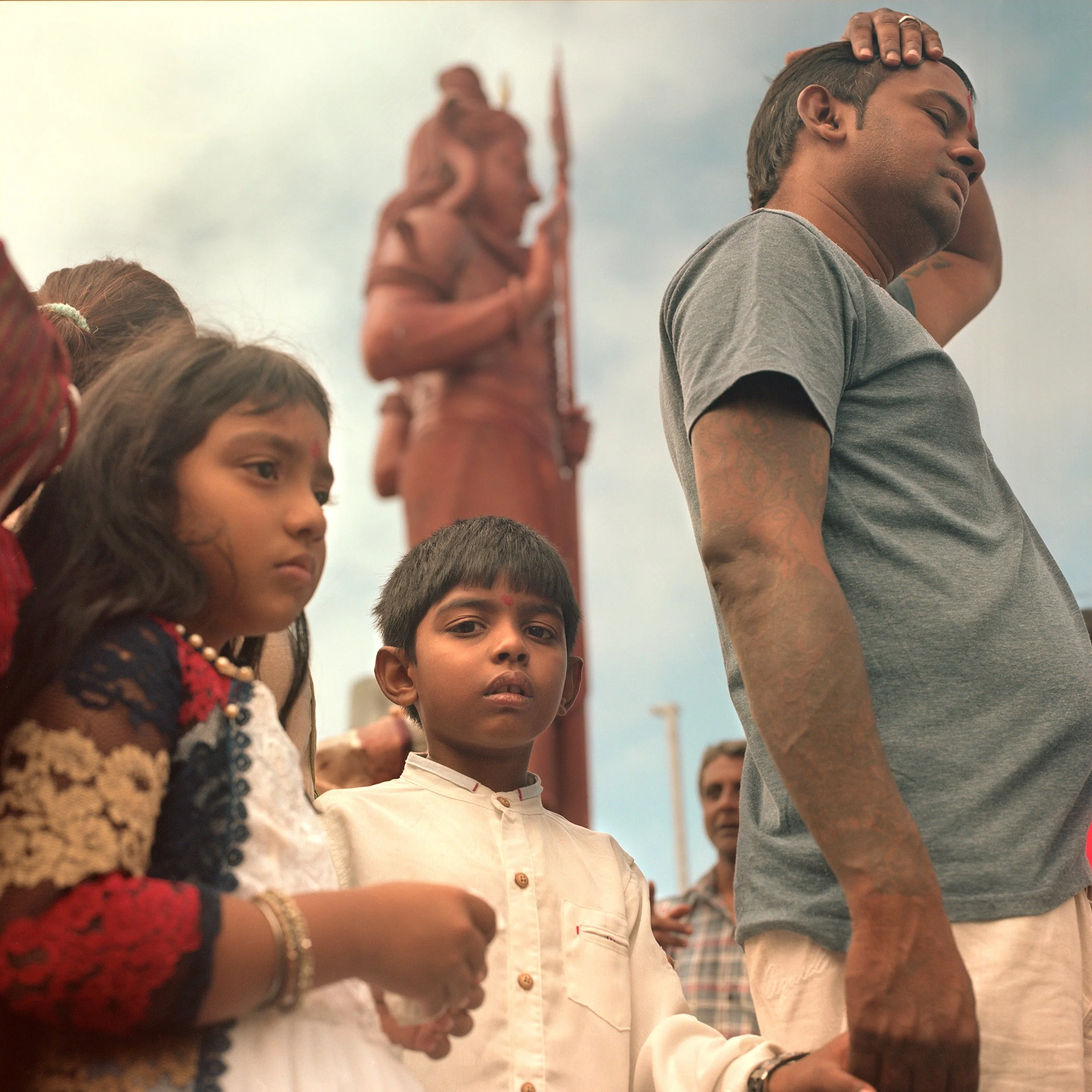 Indian man holding child's hand during a traditional event, with children and a statue of Lord Shiva in the background.