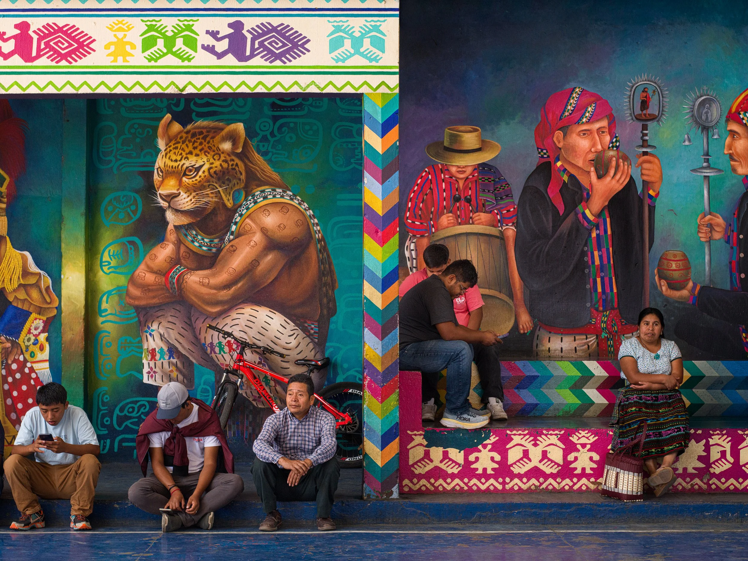 In Lake Atitlan, Guatemala, People sitting in front of colorful murals depicting indigenous figures and patterns, with one mural featuring a human figure with a leopard head.