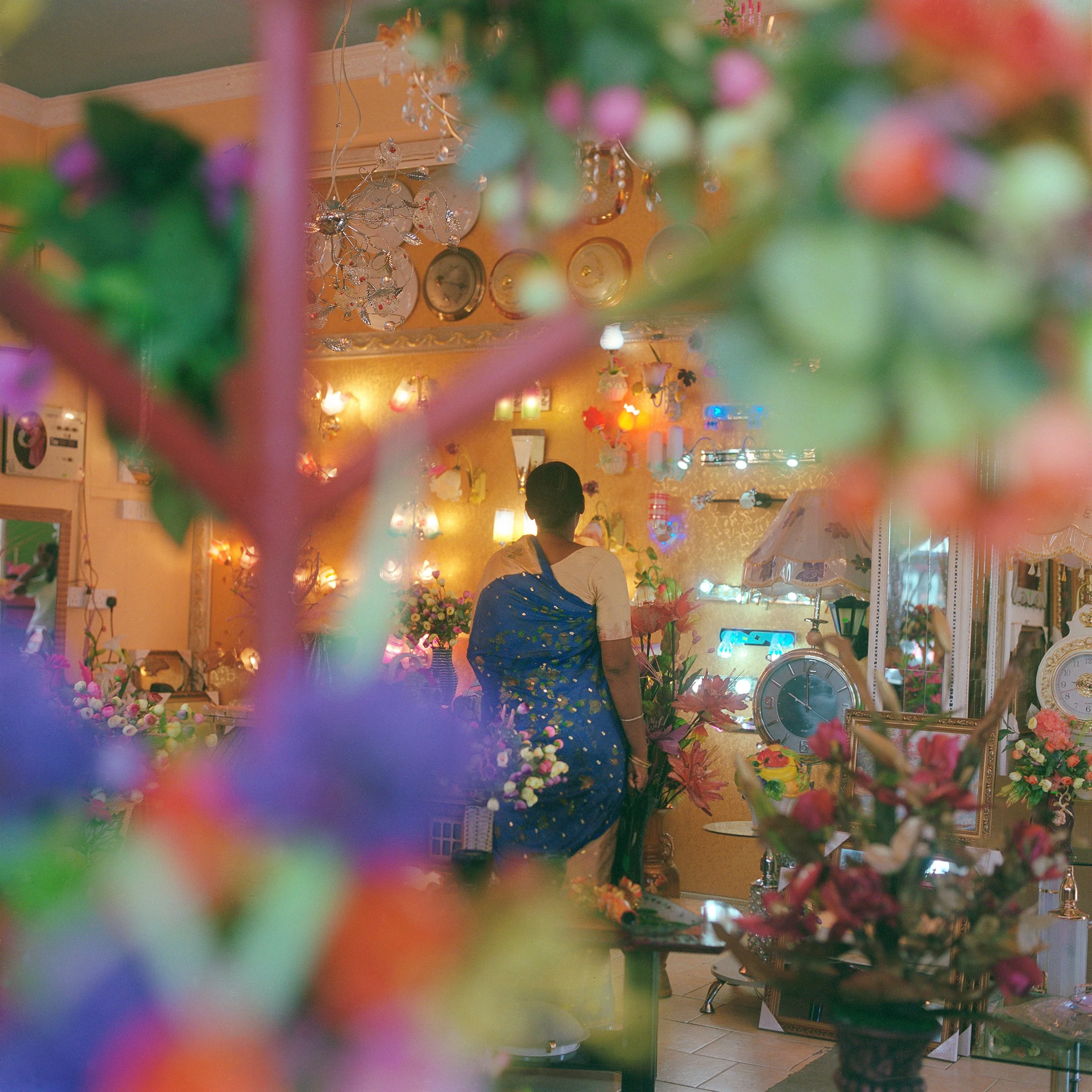 IN Mauritius, A woman in a blue saree standing in a room filled with colorful flowers, illuminated clocks, and decorative lights.
