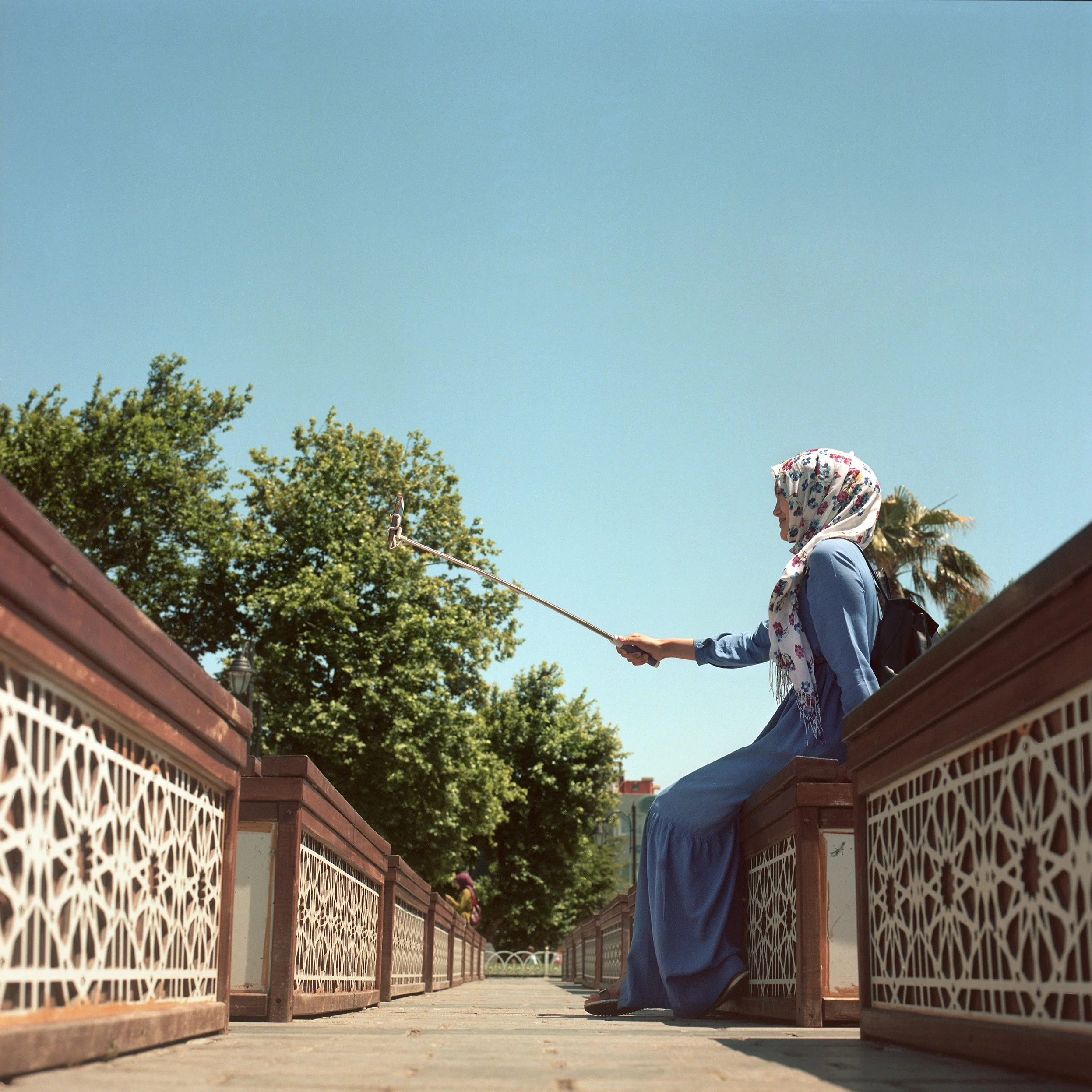 In Istanbul, Turkey, A woman wearing a floral headscarf and a blue dress sitting on a wooden bench, holding an umbrella, with trees and a person in the background under a clear blue sky.