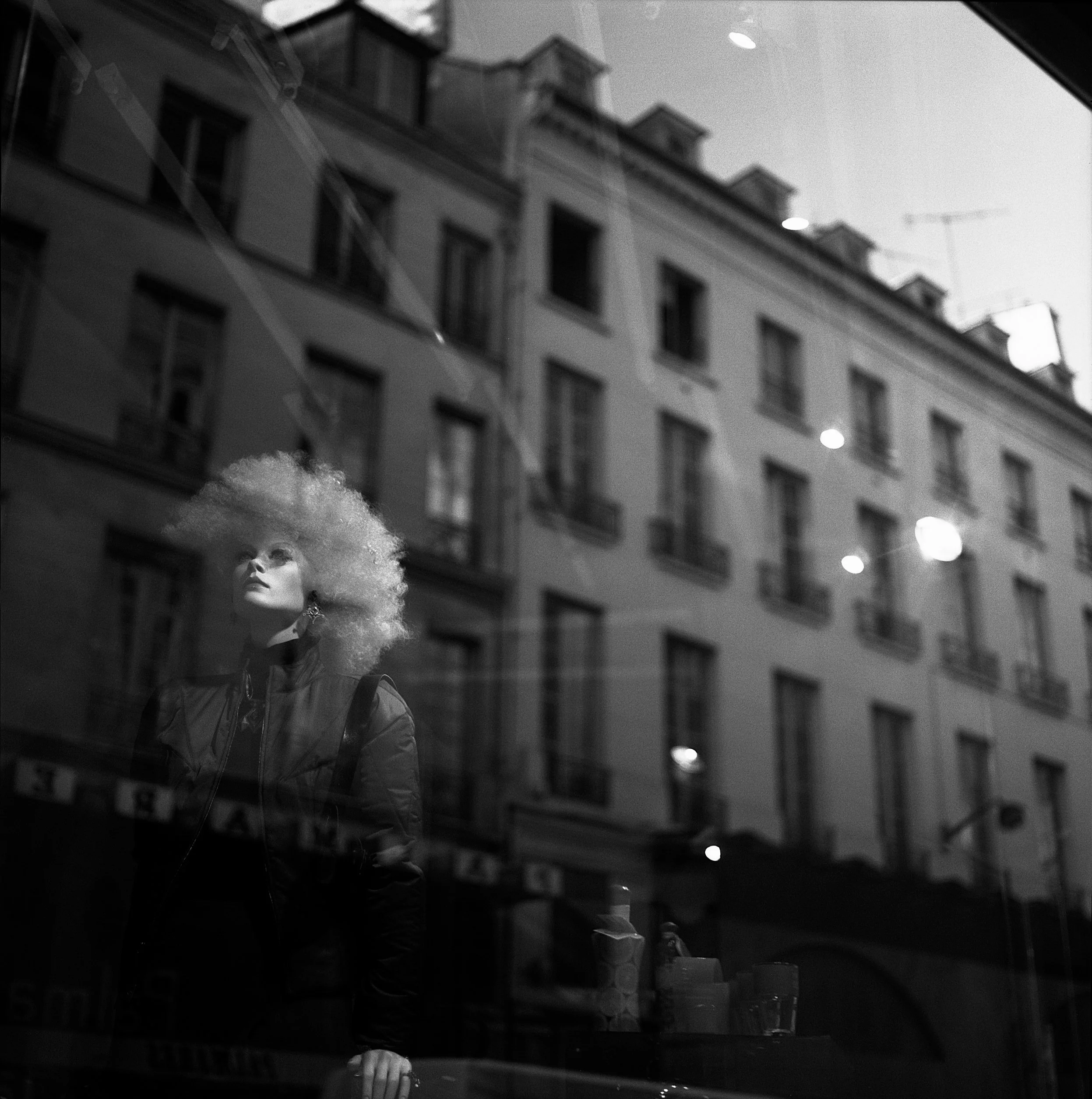 Reflection of a mannequin with curly hair and earrings in a shop window, with a building and street lights reflected in the glass.