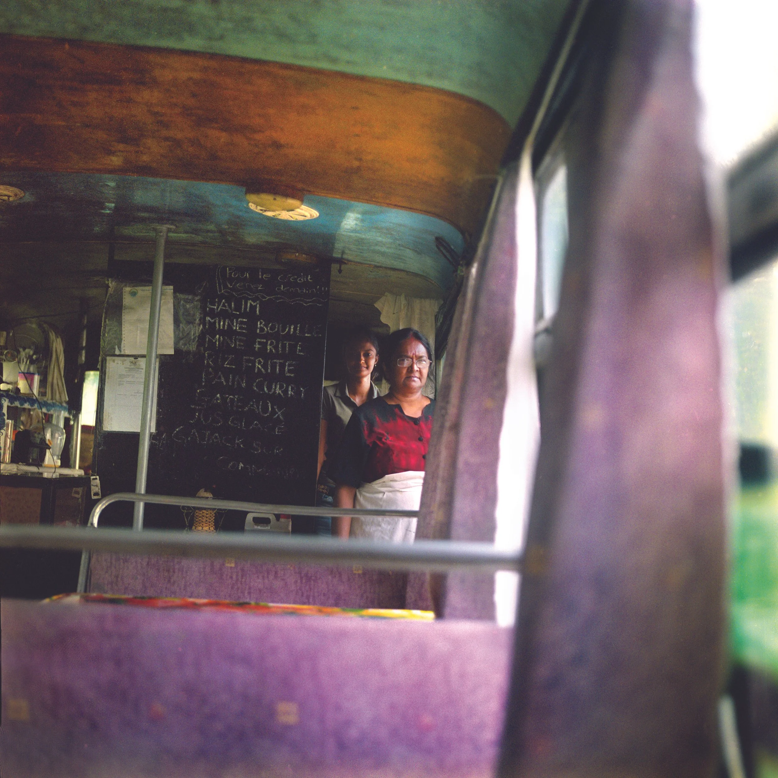 In Mauritius, Bel Ombre, Bus snack. Two women standing behind a counter inside a restaurant or cafe, with a blackboard menu behind them and various items on shelves to the side.