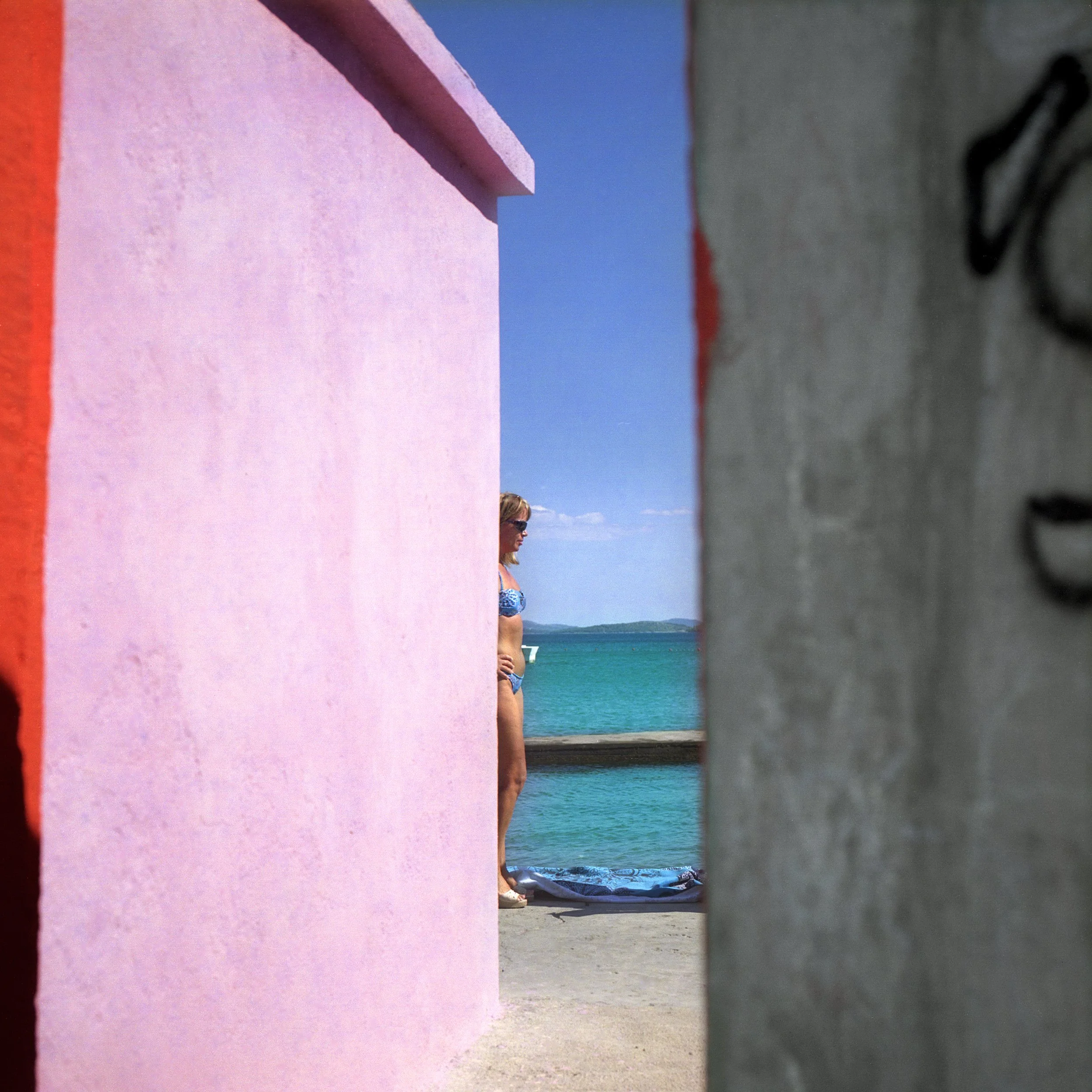 A woman standing by the water, viewed through a gap between a pink wall and a gray wall, with blue sky and distant land across the water.