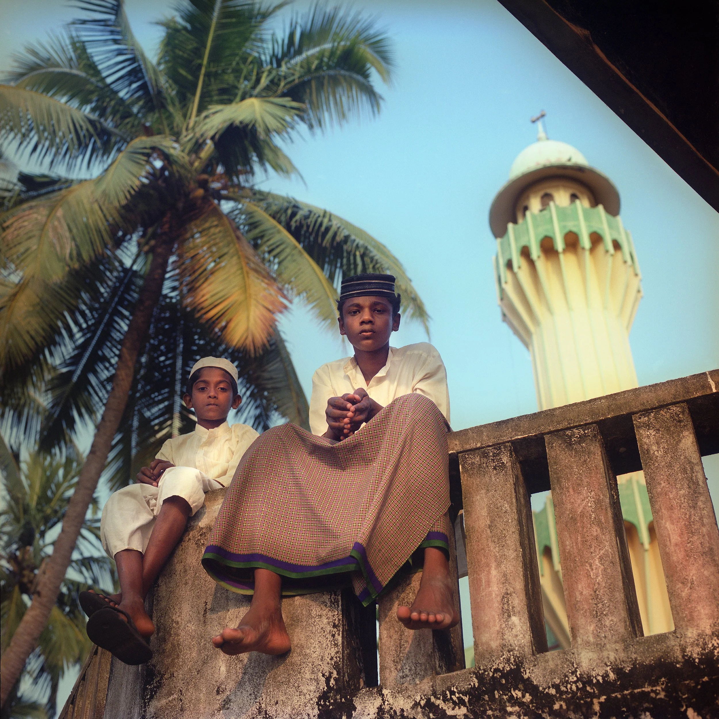 In Kerala, India, Two young boys sitting on a stone wall outdoors, with tall palm trees and a green and yellow minaret in the background, during daylight.