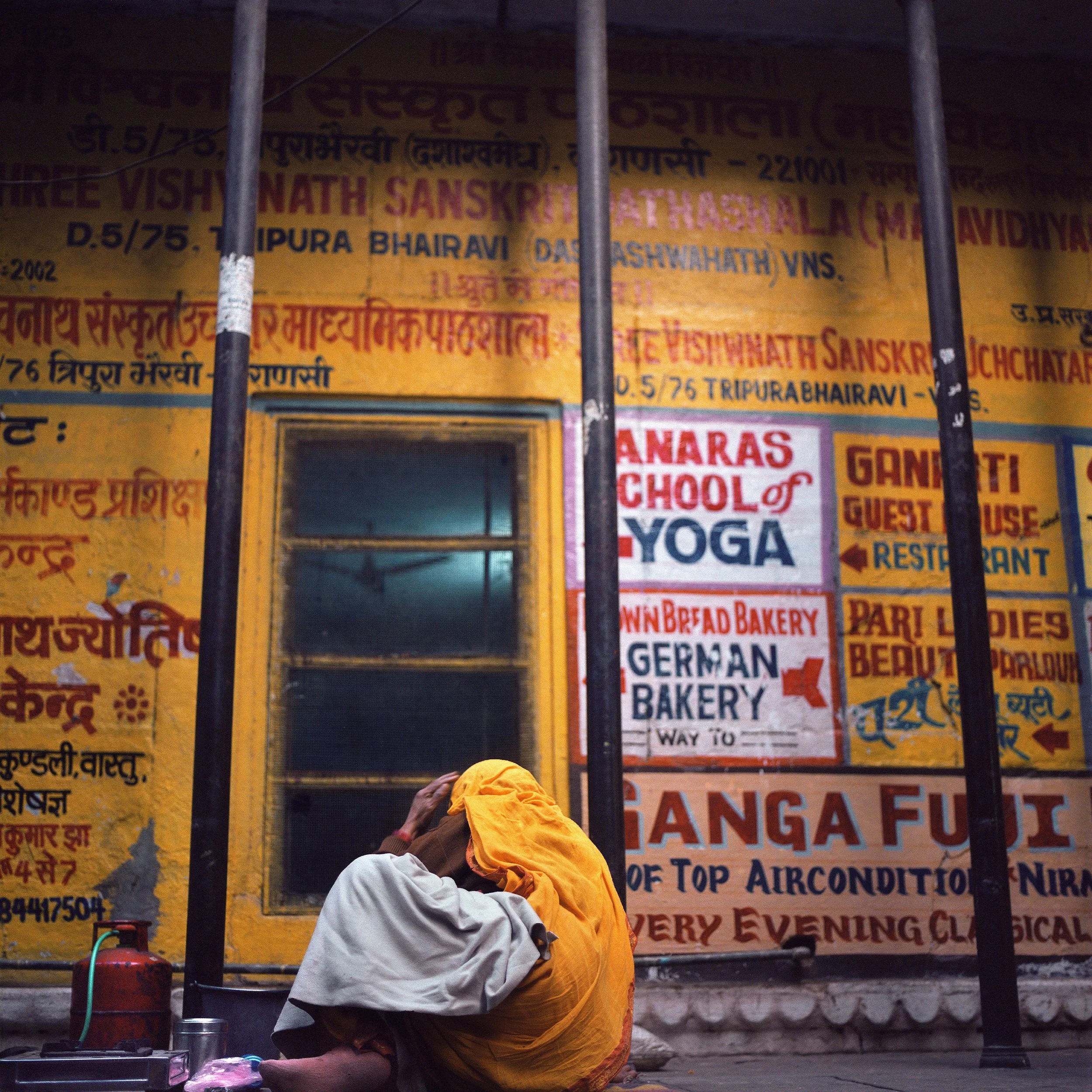 A person sitting on the ground with their head shaved, wearing a white shirt and a yellow cloth covering their face, in front of a yellow wall with colorful advertisements and text.