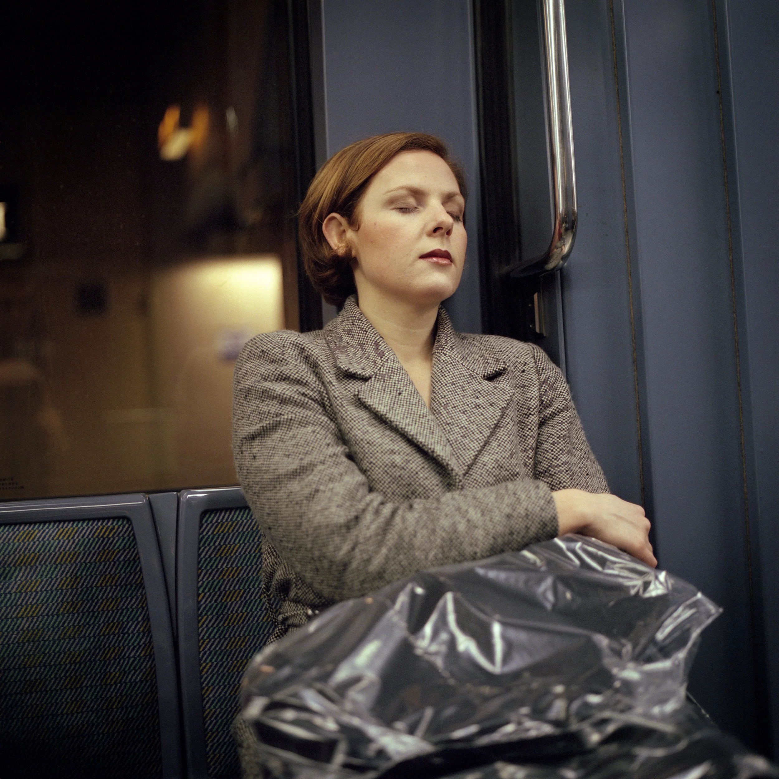 A woman with short brown hair and closed eyes sits in Paris Subway, resting with her arms crossed, wearing a gray plaid blazer. She has a serious expression and is sitting next to a window with a metal pole nearby. A plastic bag is in her lap.