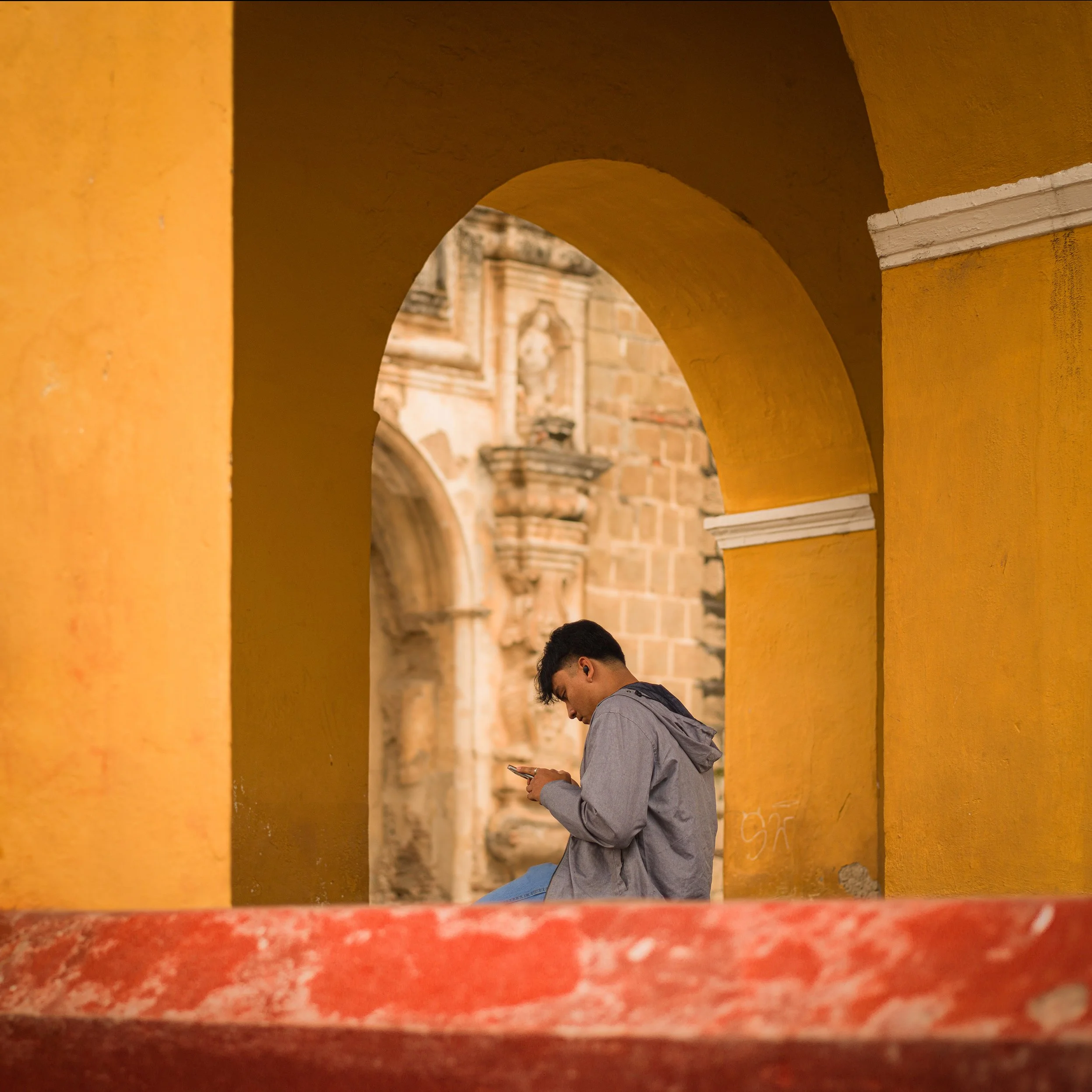 	
In Antigua, Guatemala, Young man in gray jacket seated on red ledge, looking at phone, framed by yellow arches with historic stone building in background.