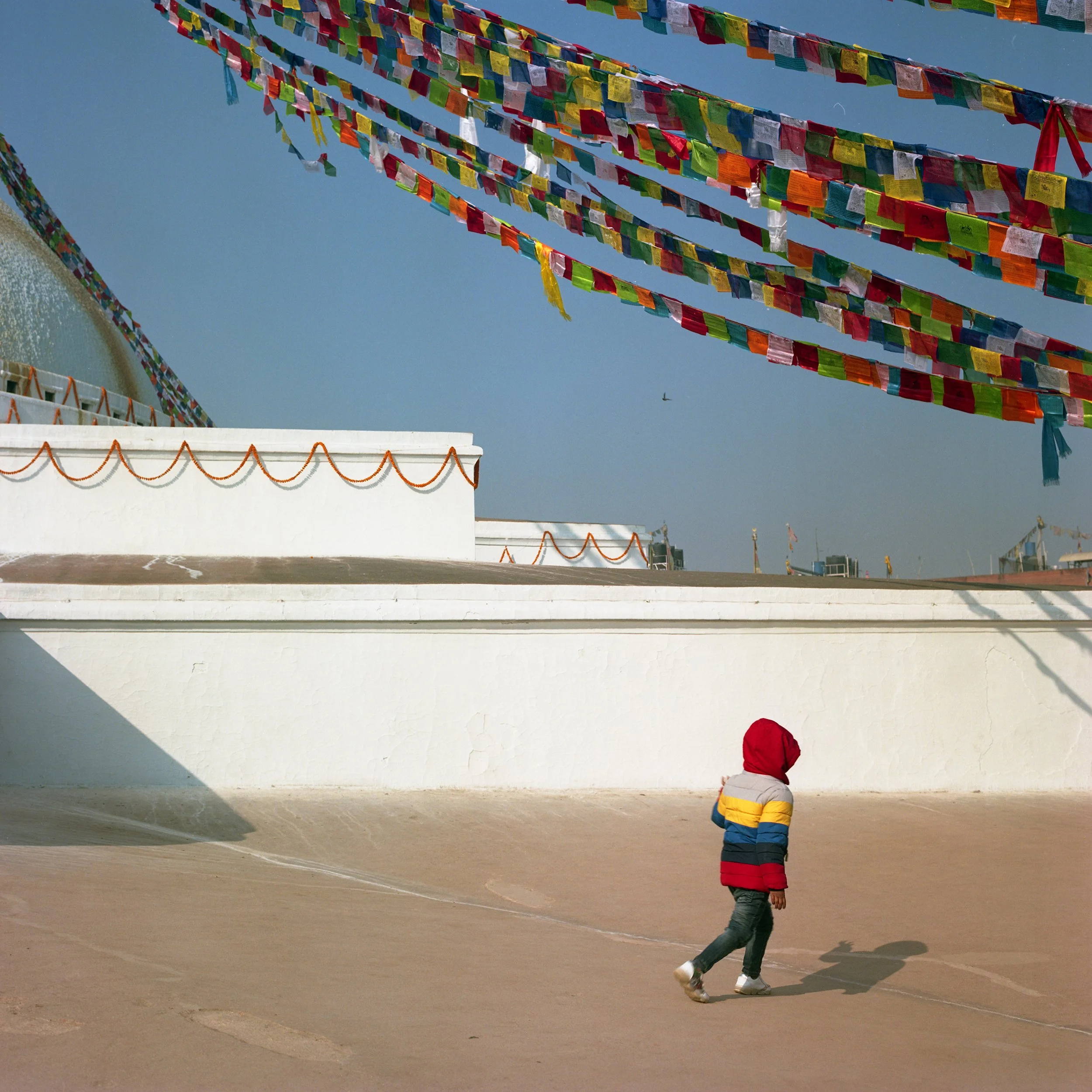 In Katmandu, A young child in a colorful, striped jacket and gray pants walking on a sandy surface with prayer flags hanging above and a white structure with orange decorations in the background, under a clear blue sky.