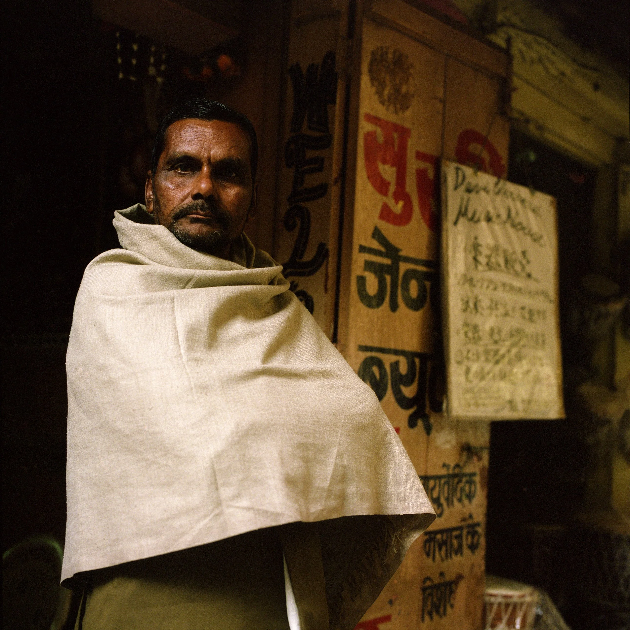 A man with dark hair and a beard, wearing a beige shawl, stands indoors in front of a wall with colorful signs and writing in Hindi.