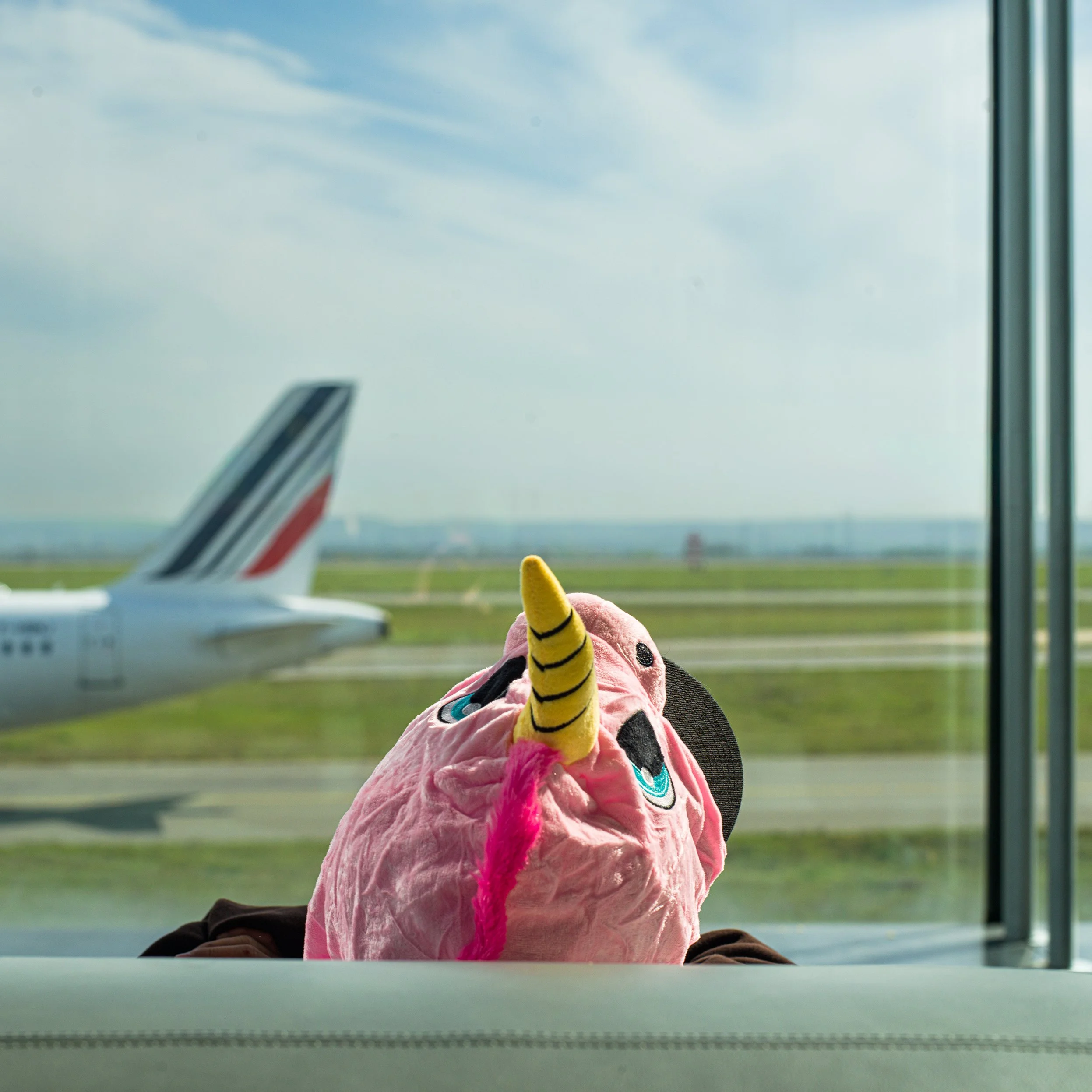 In Roissy Charles de Gaulle Airport, Paris, France, A person wearing a pink unicorn costume with a yellow horn and pink tail, sitting at an airport waiting area, looking out the window at an airplane on the runway with a clear sky.