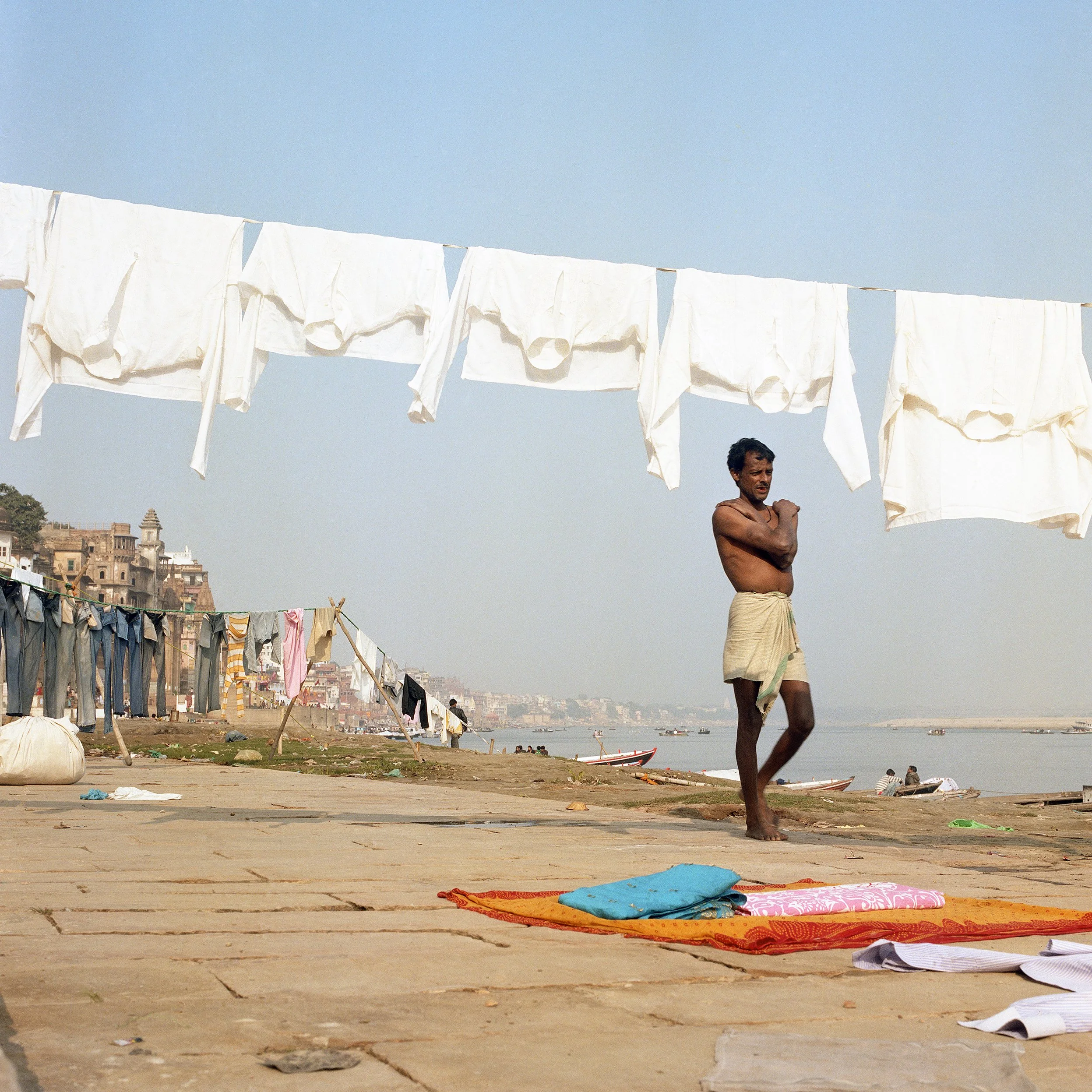 A man in traditional attire standing on a riverside pathway with white laundry hanging on a clothesline overhead, and more laundry hanging along the riverbank. The scene is set in an Indian city with buildings visible in the background.