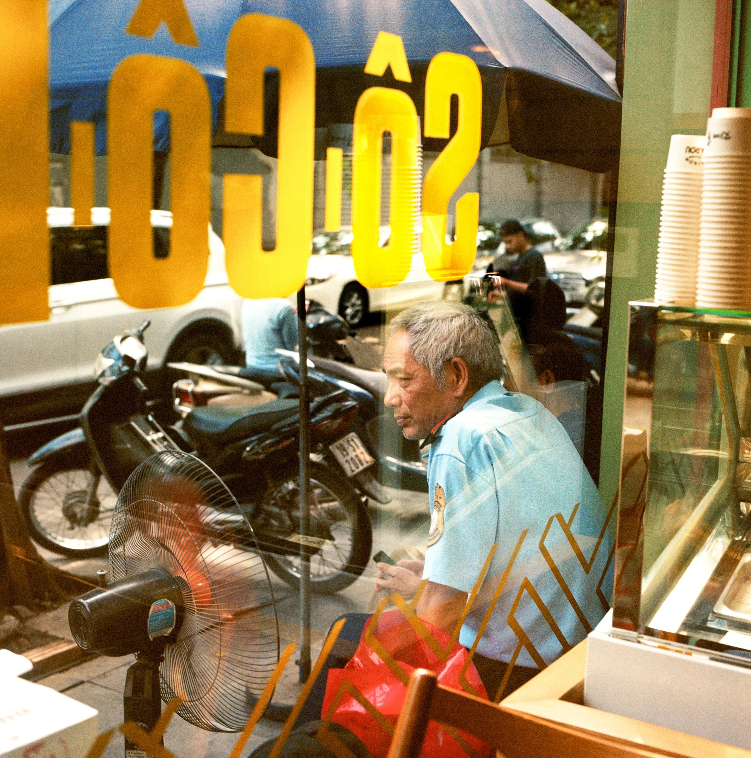 In Hanoi, Vietnam, Marou chocolat, A man in a light blue uniform sitting inside a cafe, looking at his phone, seen through a glass window with yellow numbers on it, with a street scene of parked motorcycles and cars outside.