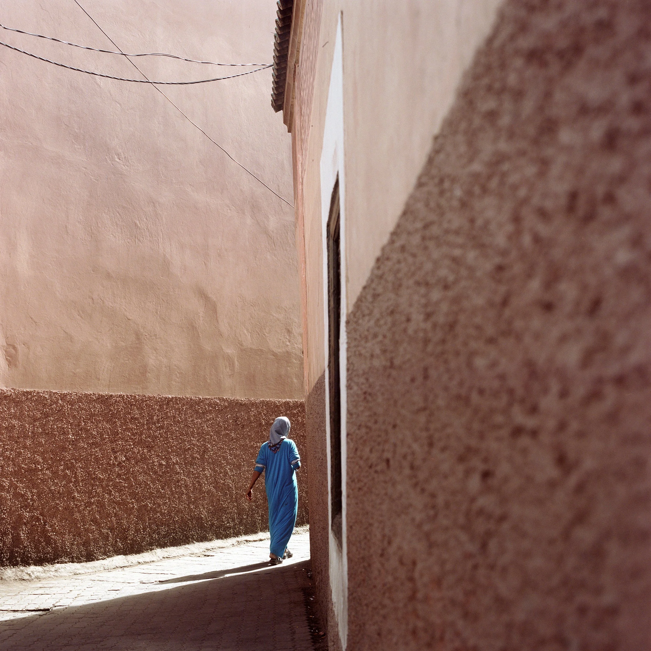 A person dressed in blue walking in a narrow alleyway between tall textured pink and beige buildings.
