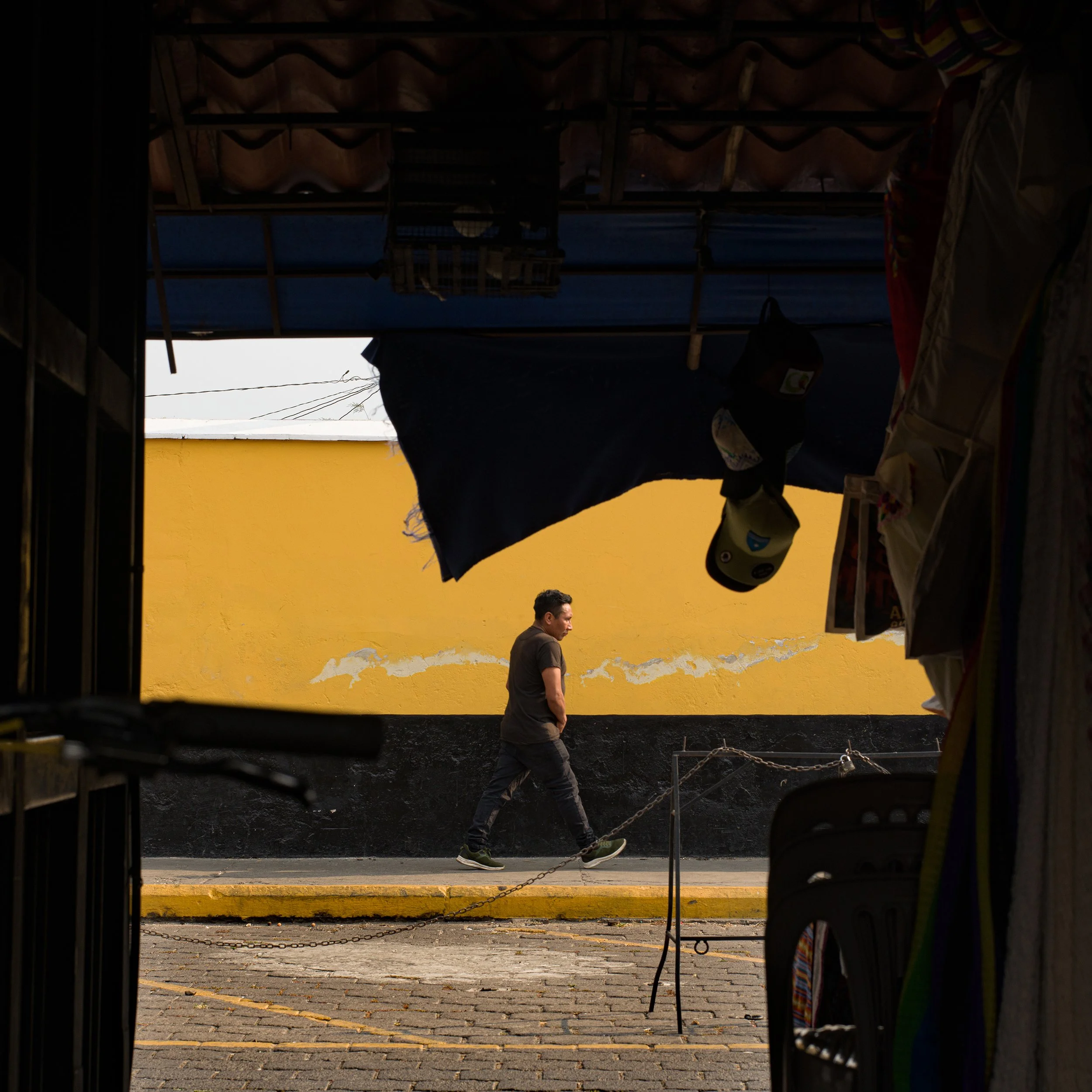 	
In Antigua, Guatemala, A man walking along a sidewalk outside, seen through an opening with hanging items and a yellow wall in the background.