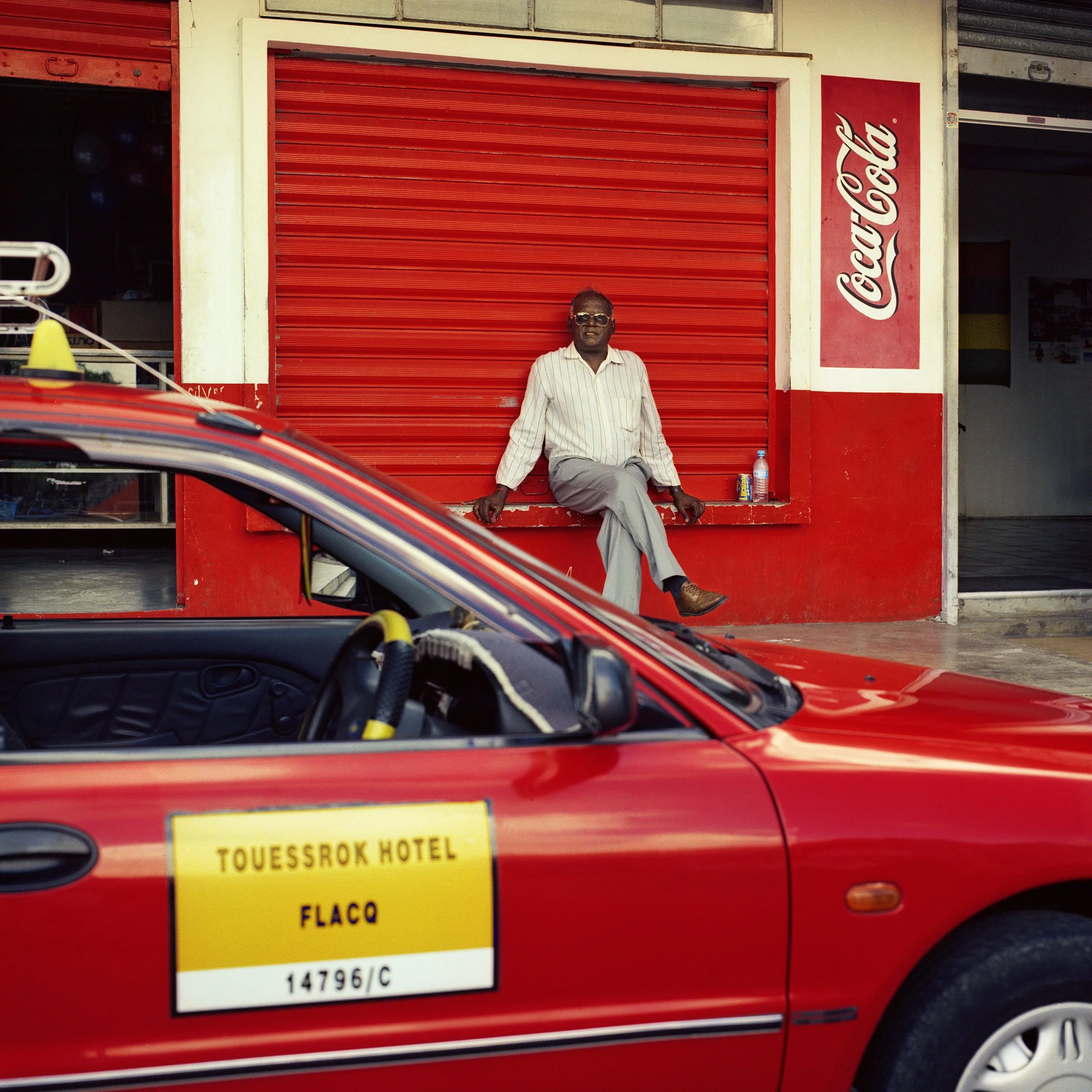 In Mauritius, Trou d'Eau Douce, A man sitting on a red ledge in front of a closed red storefront shutter with a Coca-Cola sign on the side. A red car with a yellow sign reading 'Touessrok Hotel Flaco' is parked in front.