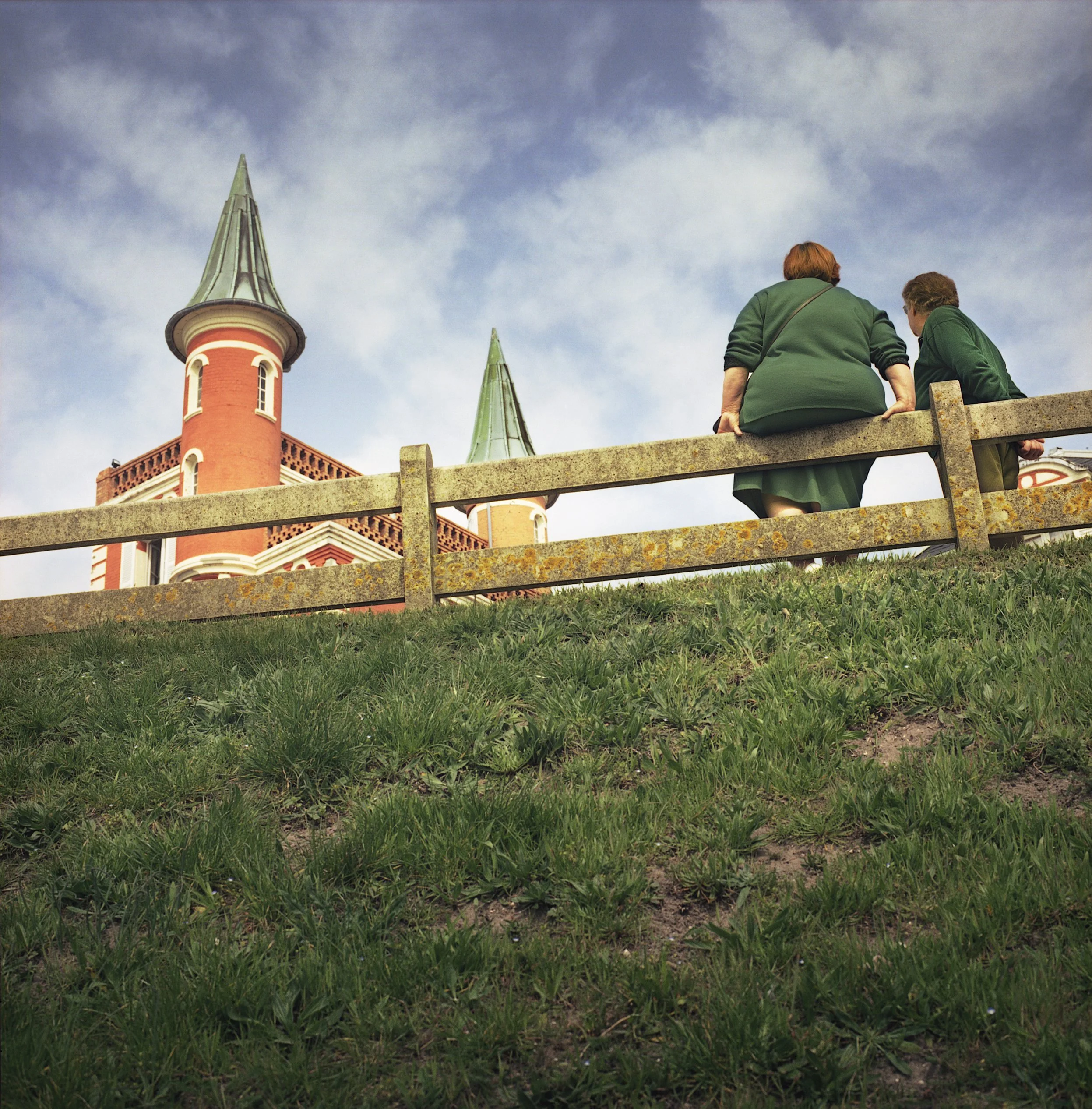 Two women in green sweatshirts sitting on a wooden fence on a grassy hill, looking towards a red brick building with tall spires under a partly cloudy sky.