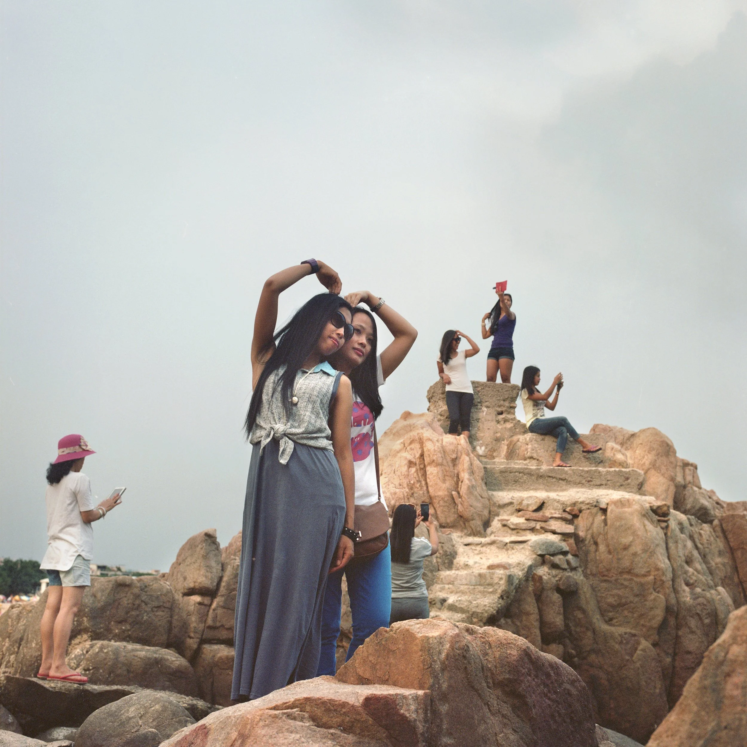 Group of women on rocky terrain, some taking photos, one standing at the top holding a red flag, others sitting or standing on rocks.