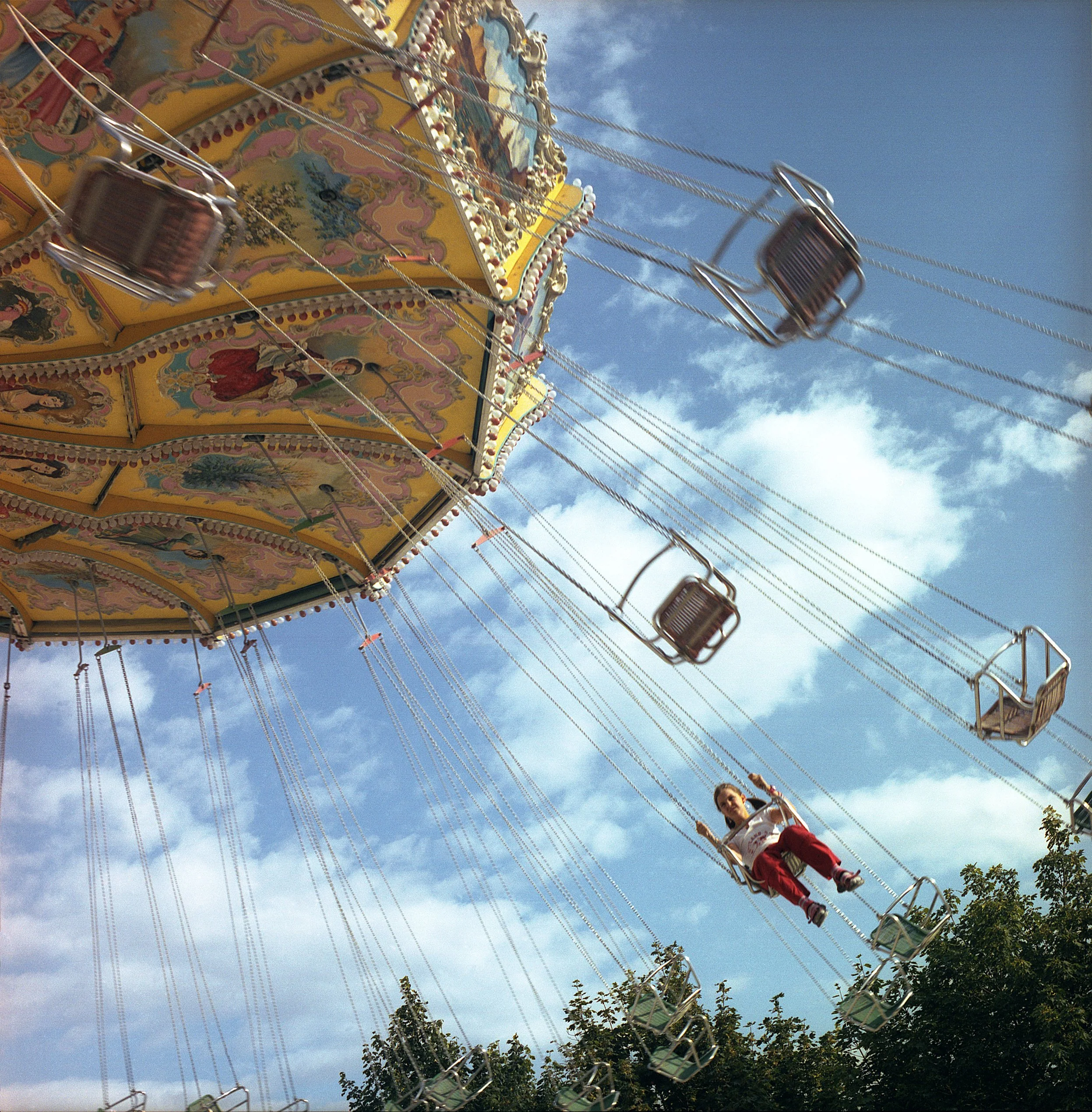 In Paris, France, A child riding on a swing ride at an amusement park during daytime, with a blue sky and some clouds in the background.
