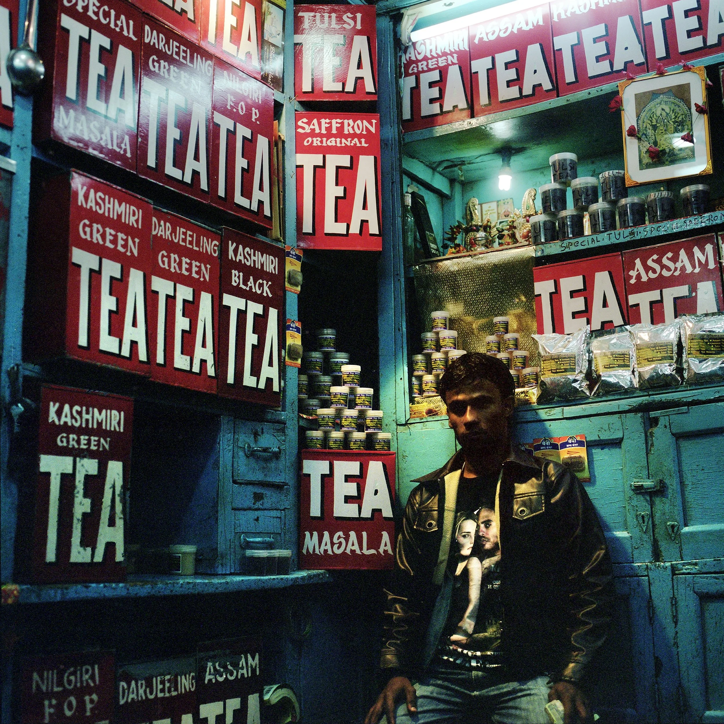 A man sitting inside a tea and spice shop with signs advertising Kashmiri, Darjeeling, Green, Black teas, and Masala tea. The shop is decorated with colorful signs and containers of spices.