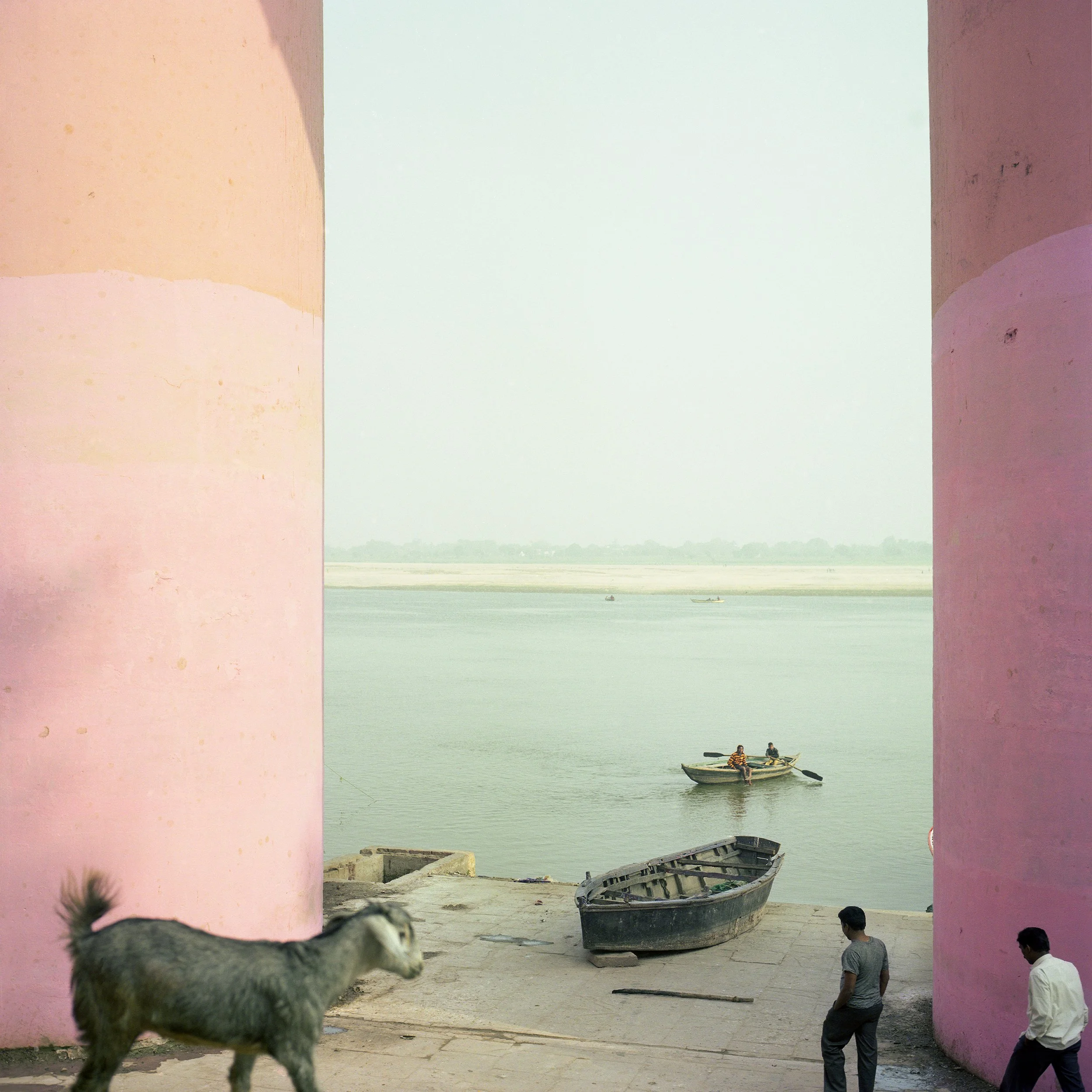 Varanassy. Benares. India. A dog in the foreground, two men walking near a pink wall, fishing boats on a river, with a hazy sky and flat land in the background.