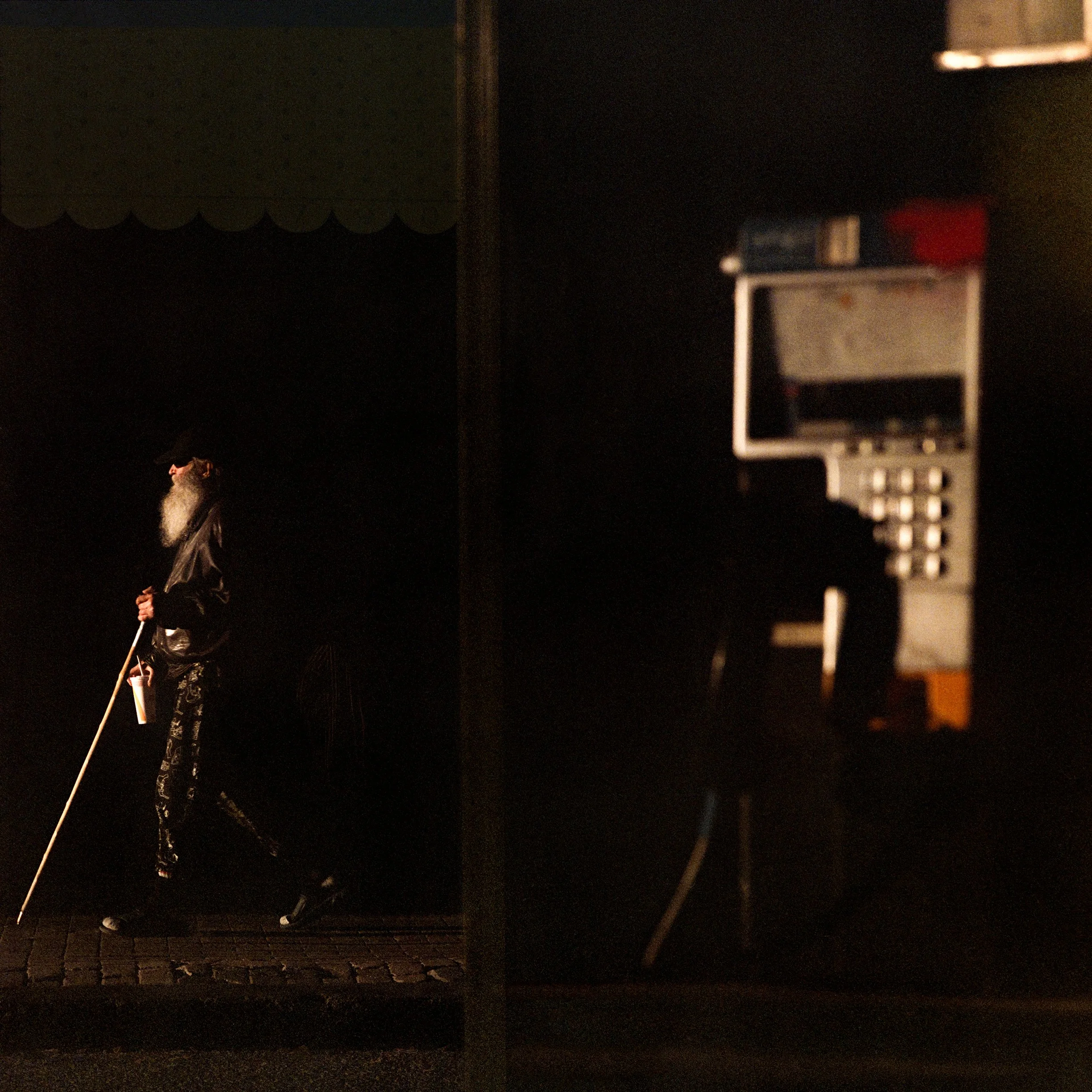 In Quebec City, Canada, A man with a long white beard and dressed in dark clothing holding a white cane, walking past a dark building at night, with a payphone visible in the foreground.
