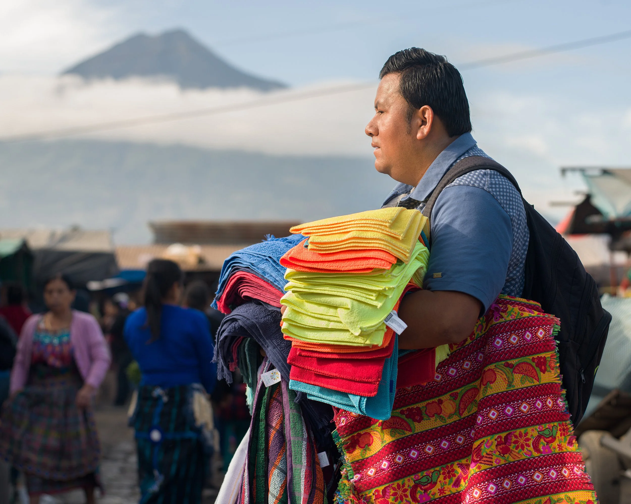In Antigua market, Guatemala, A man with a backpack holding colorful textiles, including towels and traditional woven cloth, at an outdoor market with other vendors and a mountain in the background.