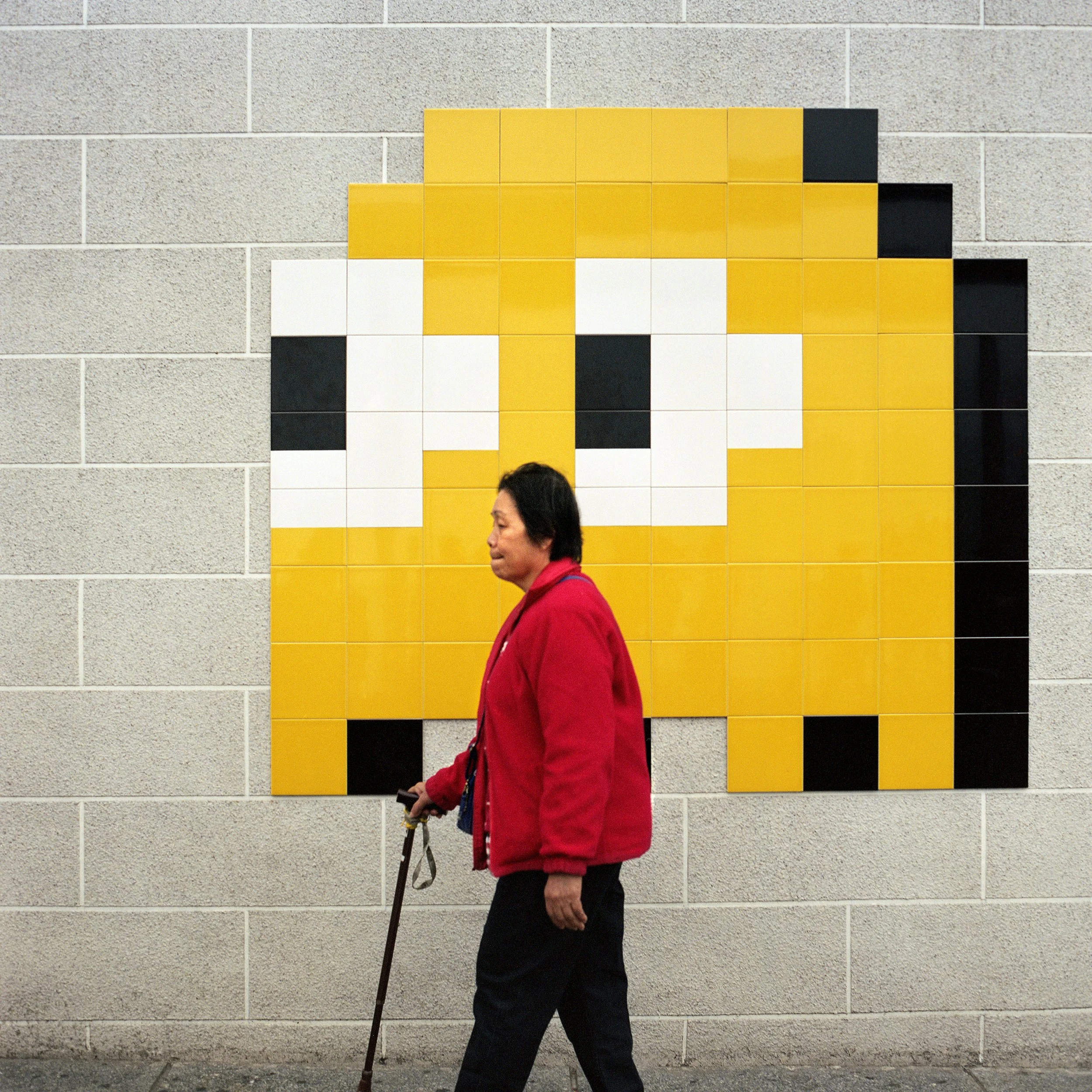 A woman in a red jacket walking past a wall-art sculpture that resembles a pixelated character, made of square tiles in yellow, black, and white.