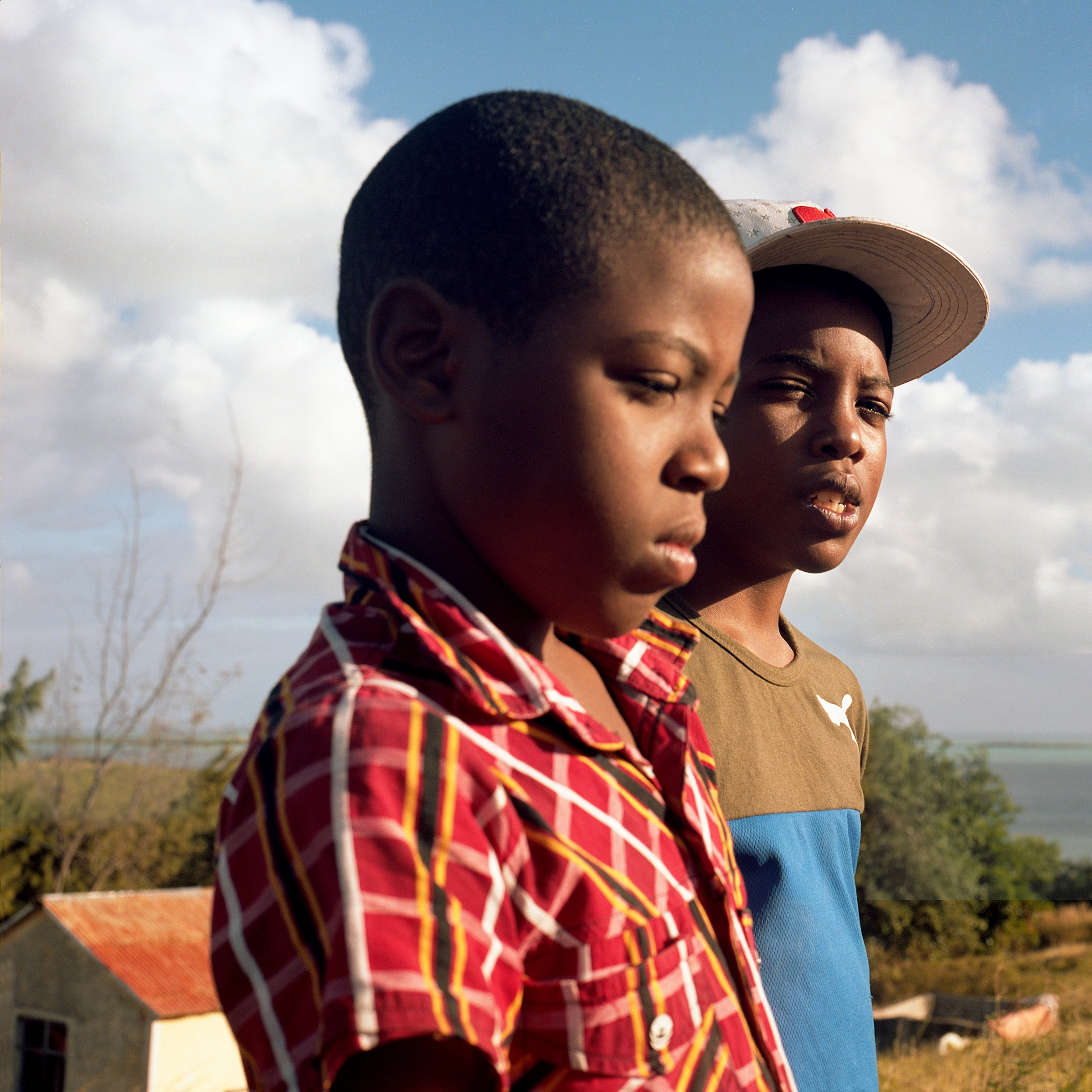 Two boys outdoors, with one in a red plaid shirt and the other in a hat and blue and brown clothing, looking thoughtfully into the distance, with houses and cloudy sky in the background.