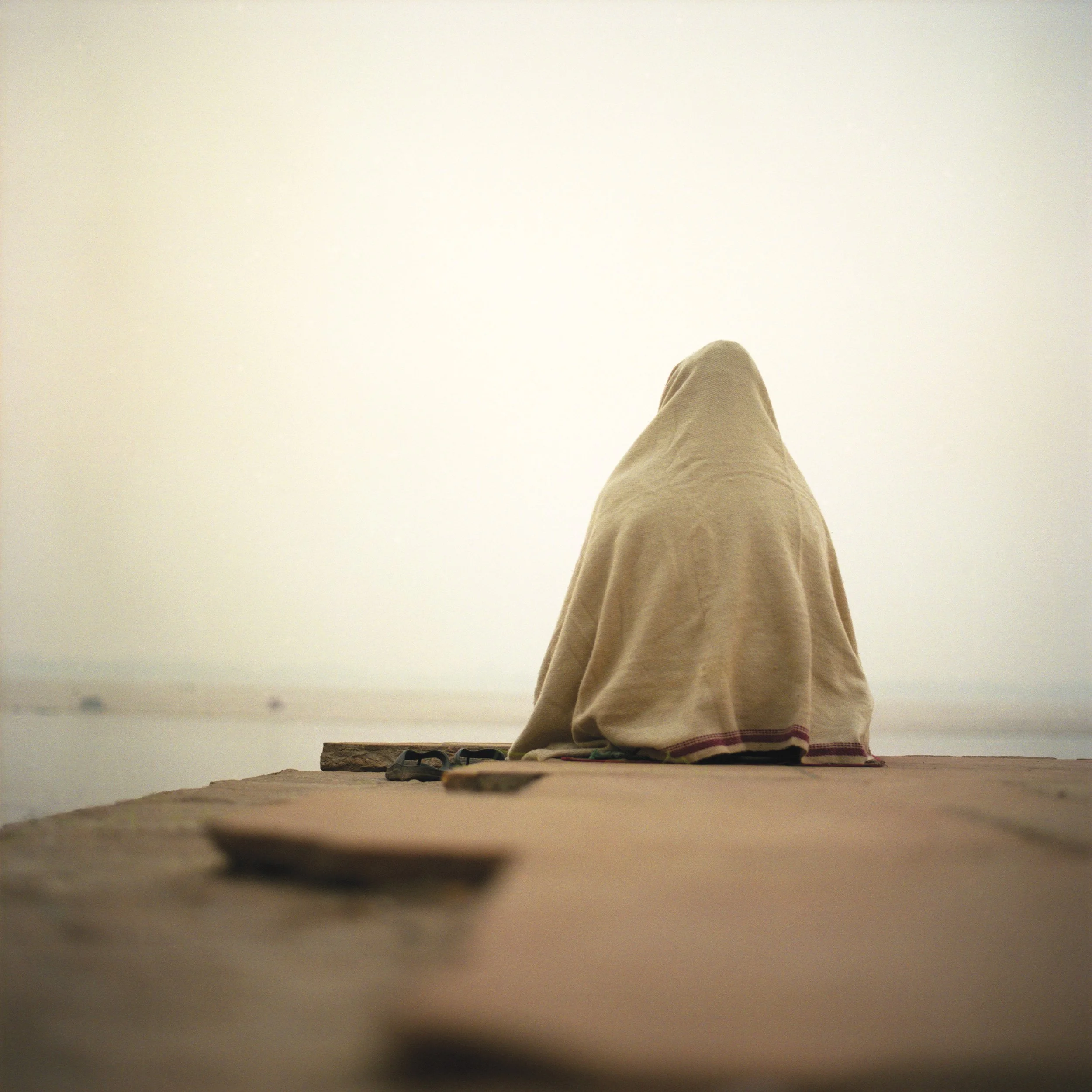 Person wrapped in a beige blanket sitting on a wooden deck overlooking a body of water, with a hazy sky in the background.