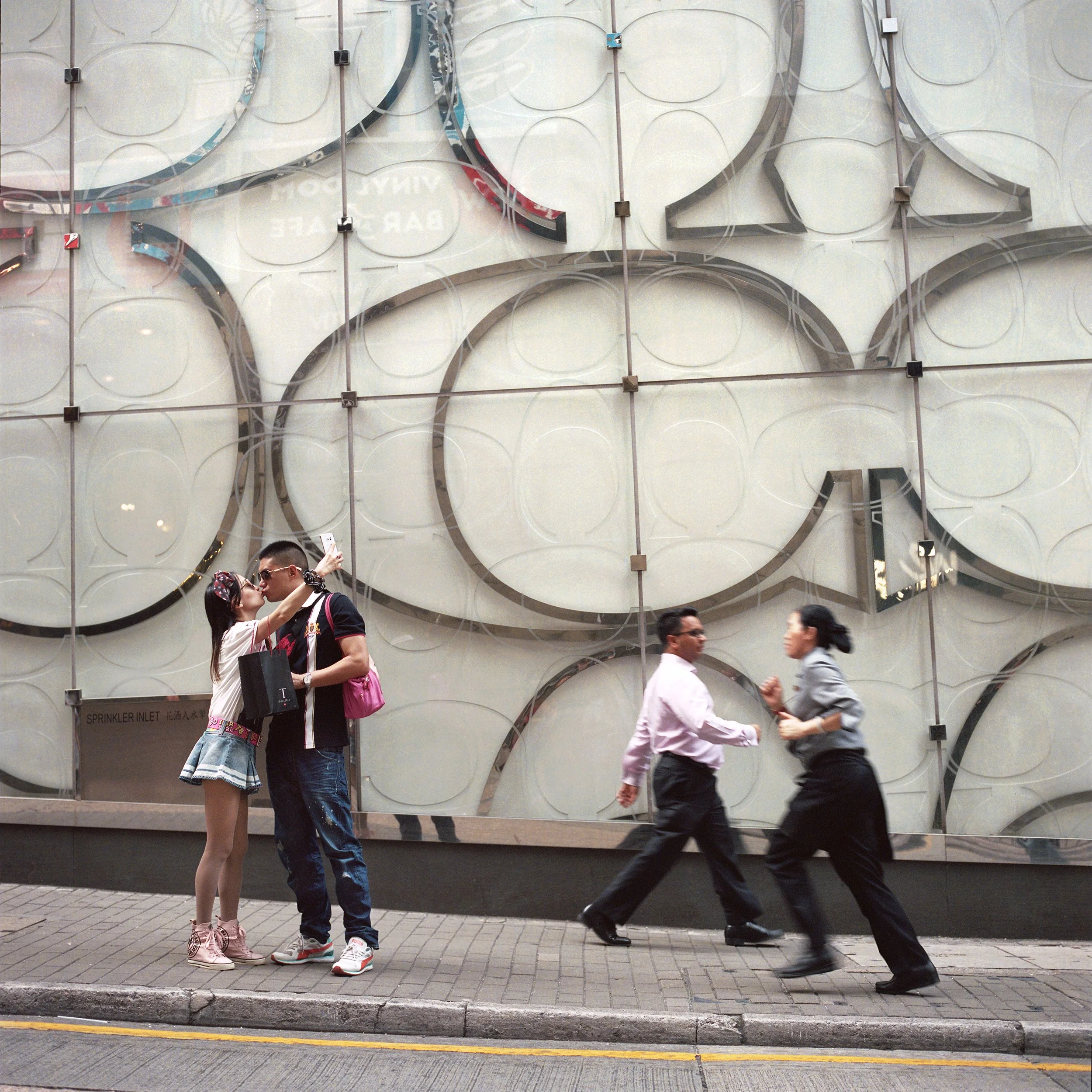 Street scene with two couples walking and a couple taking a photo of each other in front of a large modern building with geometric circular designs.