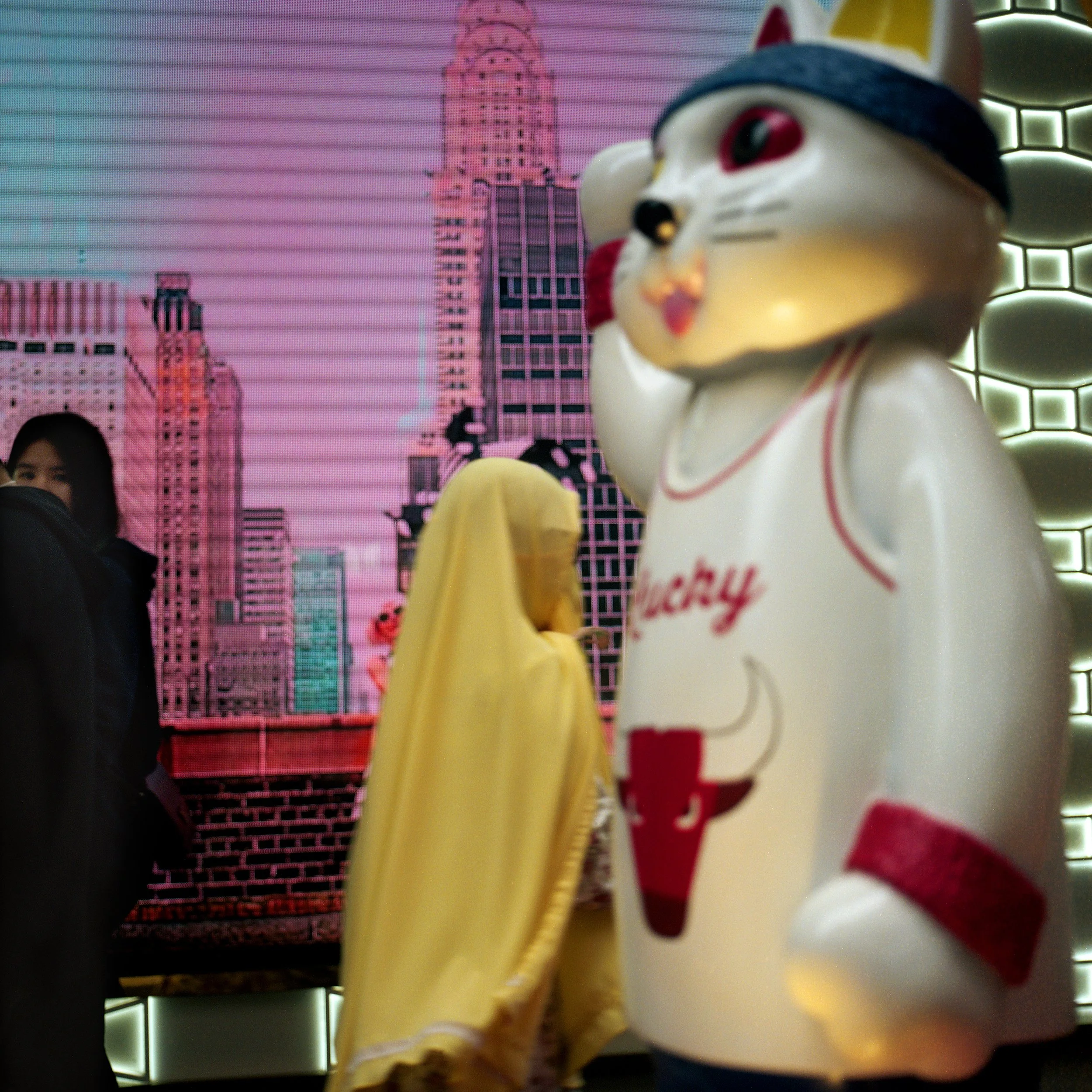 Close-up of a large Lucky Cat statue wearing a baseball cap and a shirt with a drink illustration, in front of a pink cityscape mural.