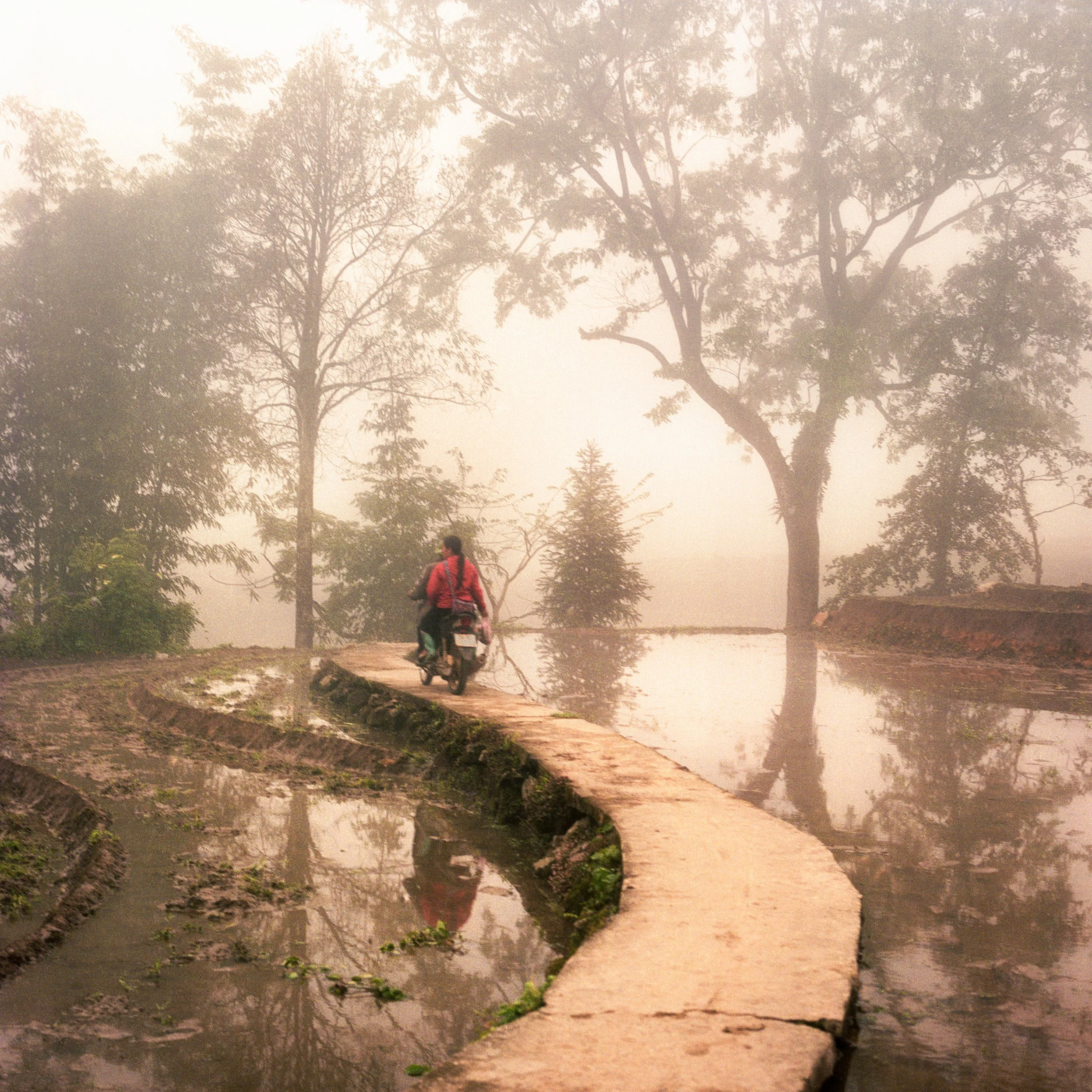 In Sa Pa, Vietnam, A person riding a motorcycle along a narrow concrete path through a flooded area with trees in the background and foggy weather.